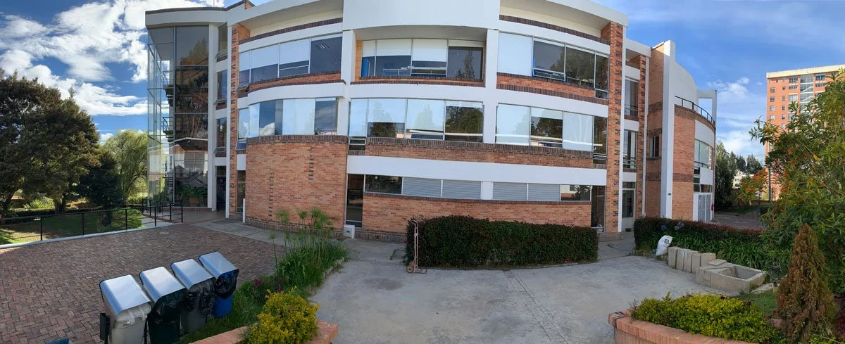 Panoramic view of a modern brick and glass apartment building with multiple large windows, surrounded by greenery and clear blue sky.