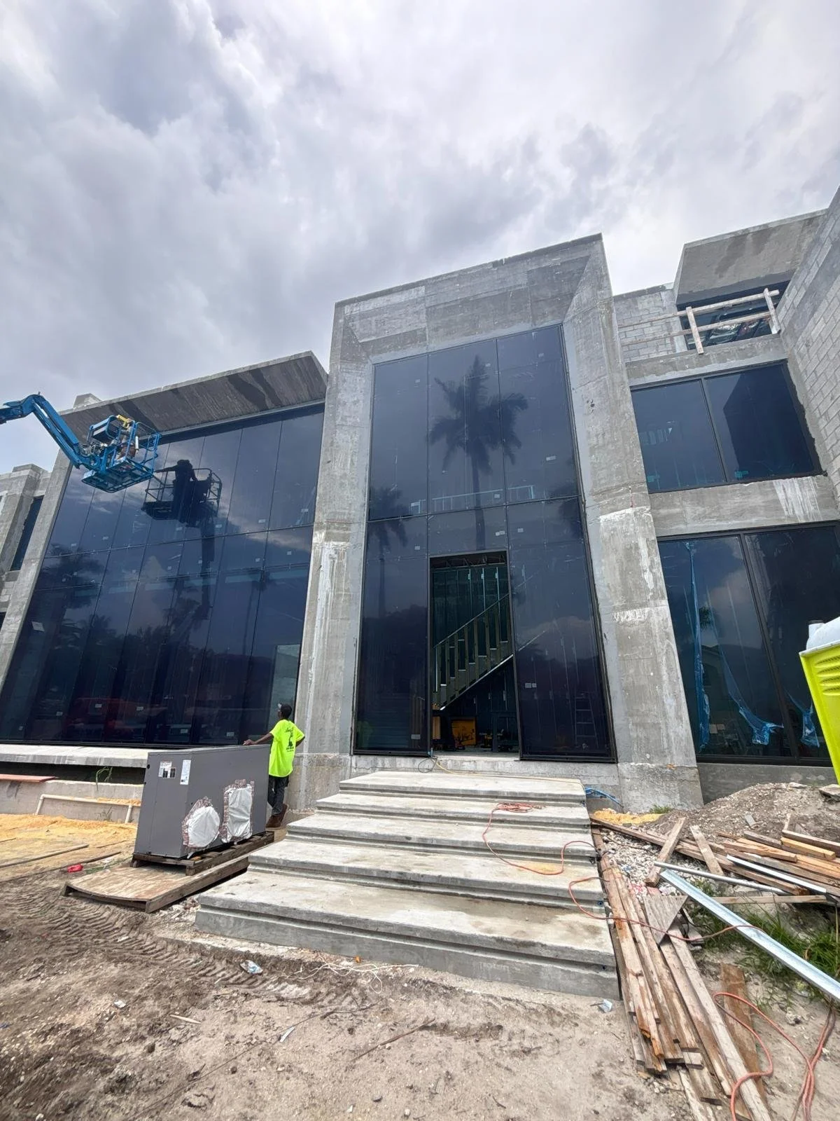 Construction site of a modern building with large glass windows, concrete structure, and a staircase. A worker in a green shirt is near electrical equipment, and a lift is working on the upper part of the building.