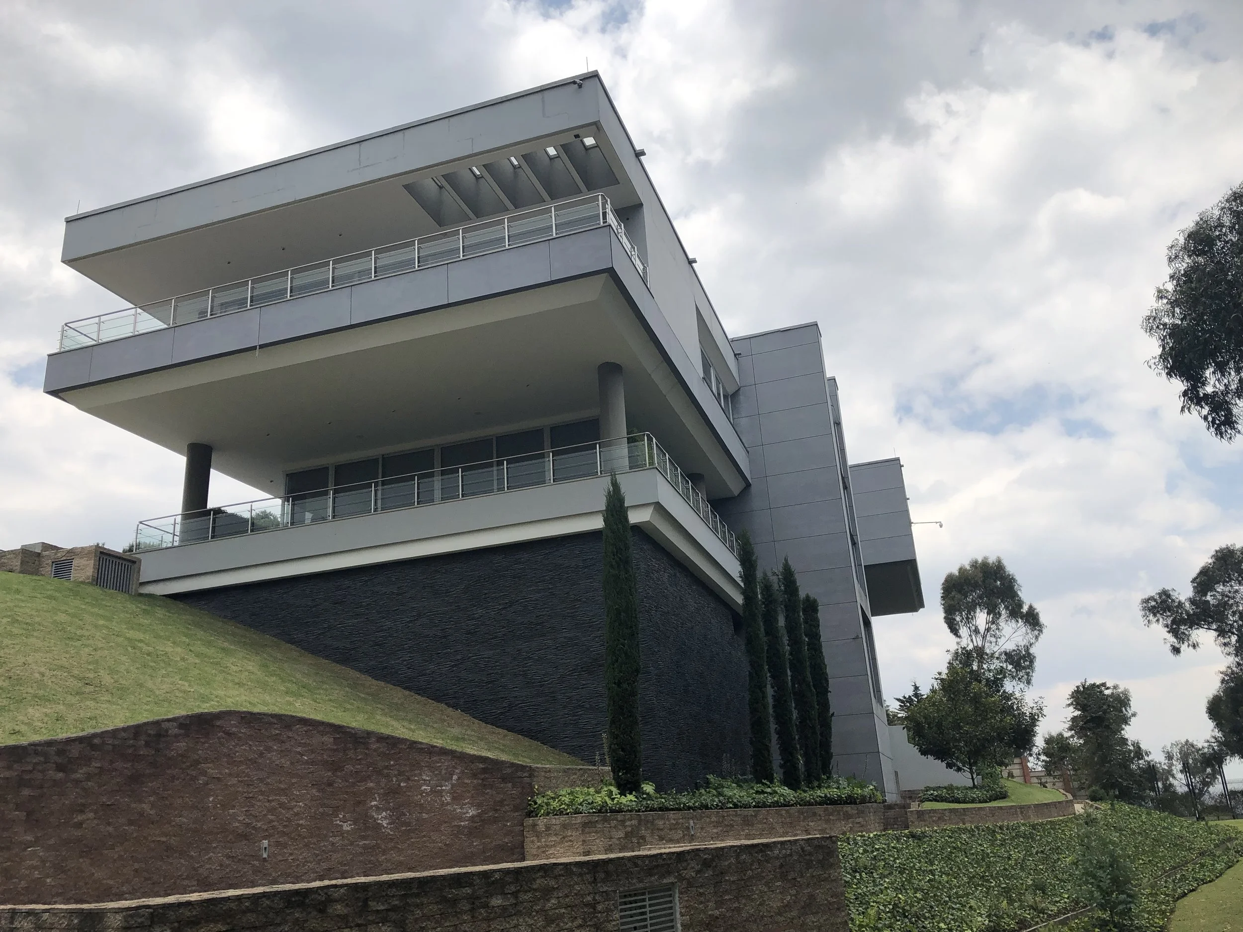 Modern multi-story building with large balconies, supported by columns, set on a grassy hill with trees and landscaped plants.