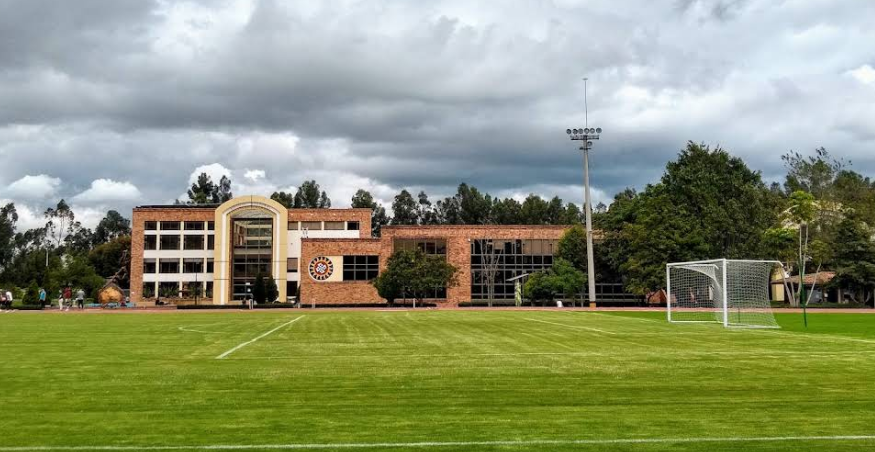 A large grassy sports field with a soccer goal, surrounded by trees and a brick building in the background, under a partly cloudy sky.