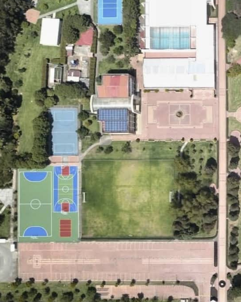 Aerial view of a sports complex featuring basketball courts, tennis courts, a soccer field, and indoor swimming pools surrounded by trees and pathways.