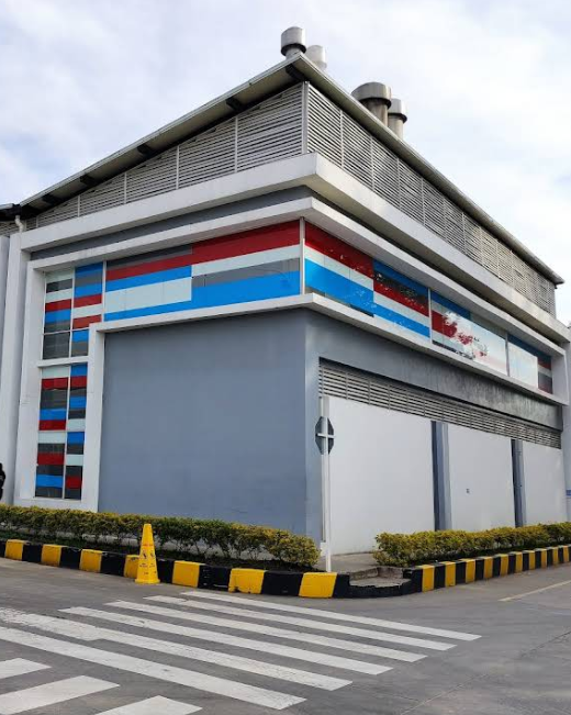 A modern two-story building with gray walls, decorated with red, blue, and white horizontal stripes on the upper part. It sits next to a crosswalk and a parking lot, with yellow and black curb barriers and small green bushes along the sidewalk.