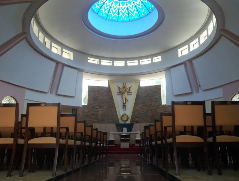 Inside view of a church with wooden chairs arranged in rows, an altar at the front, and a crucifix behind it. The ceiling features a large stained glass skylight.