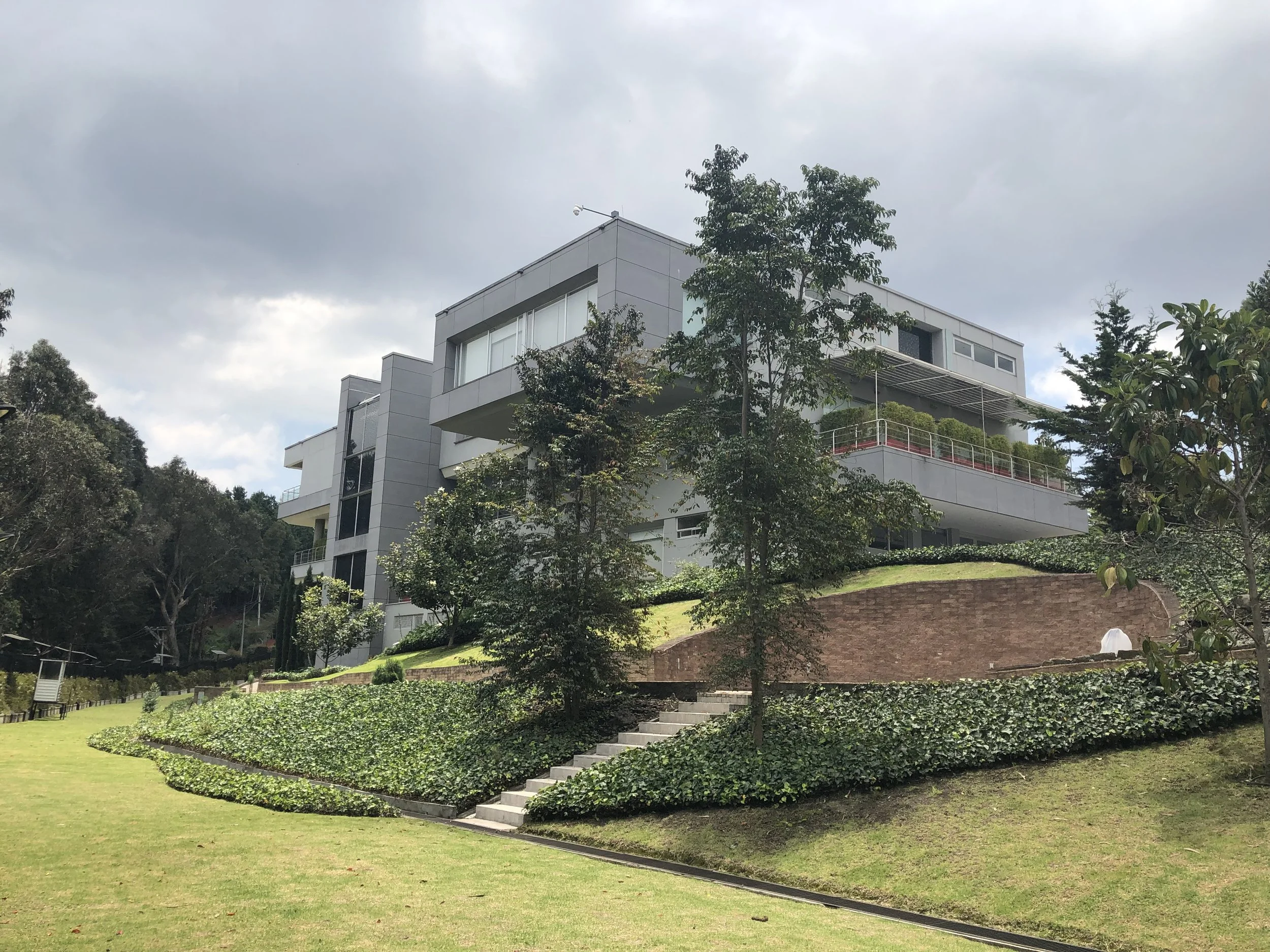 Modern multi-story residential building with large windows, surrounded by green landscaping and trees, under a cloudy sky.