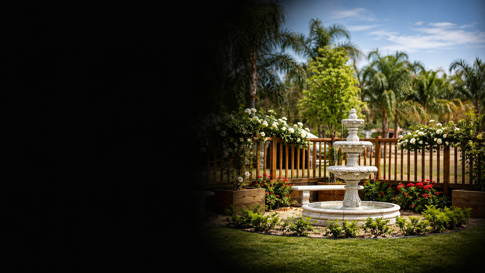 A white three-tier garden fountain surrounded by flowering plants and a wooden fence, with palm trees and a partly cloudy sky in the background.