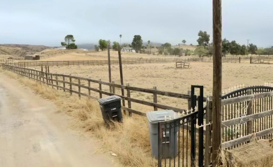 A sandy rural field enclosed by a wooden fence with a black metal gate, two trash bins near the gate, and utility poles, with sparse trees and buildings in the distance under an overcast sky.