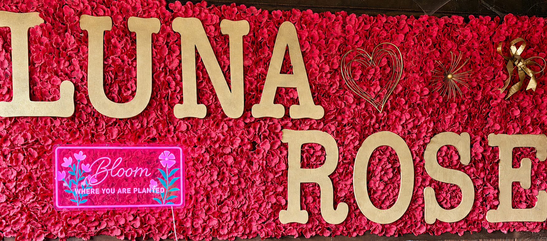 Decorative display with pink flower wall background, large beige letters spelling 'LUNA' and 'ROSE', and a neon sign that says 'Bloom WHERE YOU ARE PLANTED' with flower illustrations.