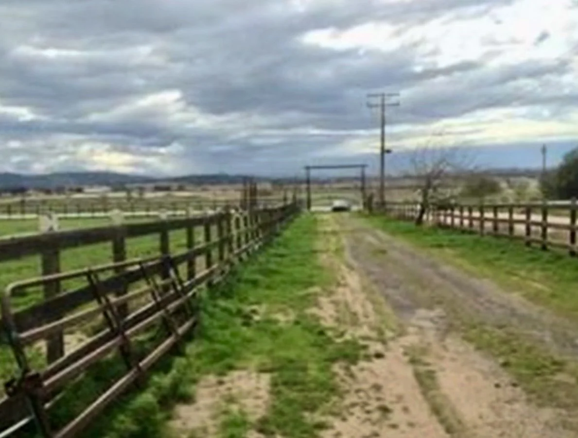 A dirt path flanked by wooden fences on either side, with a cloudy sky overhead and power lines in the background.