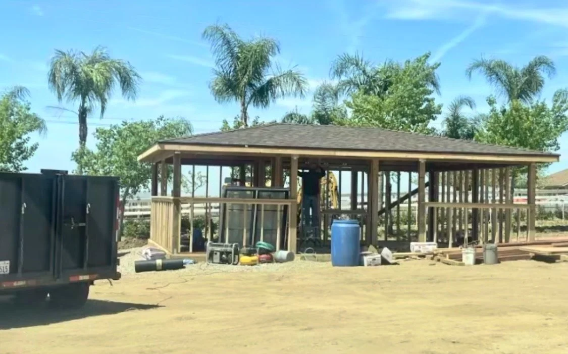 Construction site with a wooden structure being built, surrounded by construction tools and equipment, with palm trees and clear blue sky in the background.