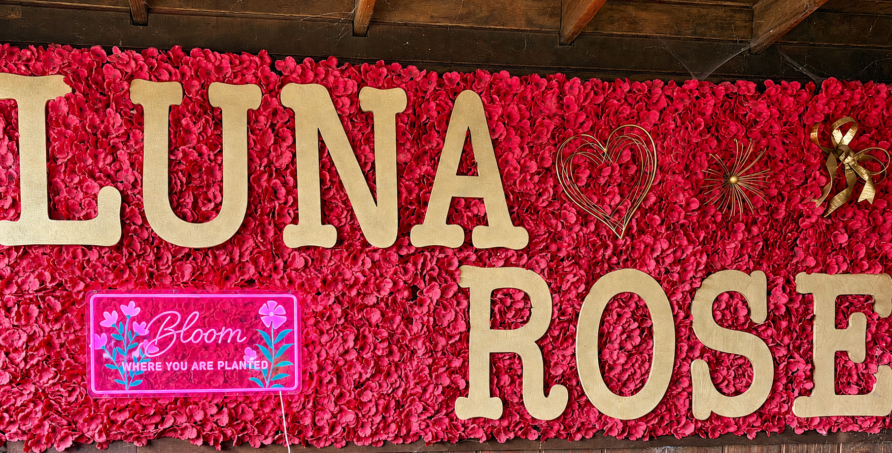 A wall decorated with pink flower petals, with large gold letters spelling 'LUNA' and 'ROSE,' along with decorative gold hearts and stars. A neon pink sign reads 'Bloom WHERE YOU ARE PLANTED' with floral illustrations.