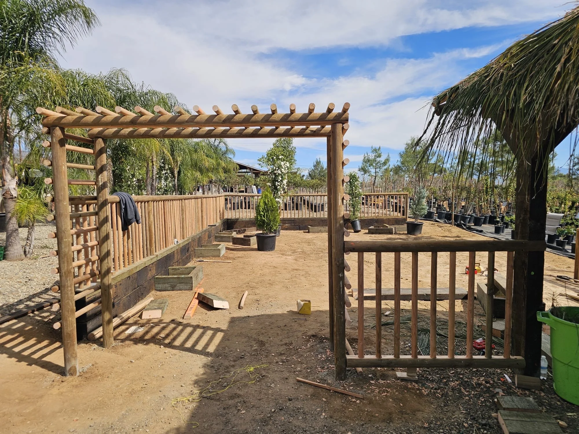 A wooden garden archway under construction with a ladder detail, fenced in an area with potted trees and plants, palm trees, and a partly cloudy blue sky.