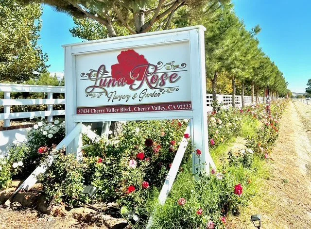 A white sign for Luna Rose Nursery & Garden with a red rose graphic, located at 37434 Cherry Valley Blvd, Cherry Valley, California, surrounded by blooming flowers and trees.