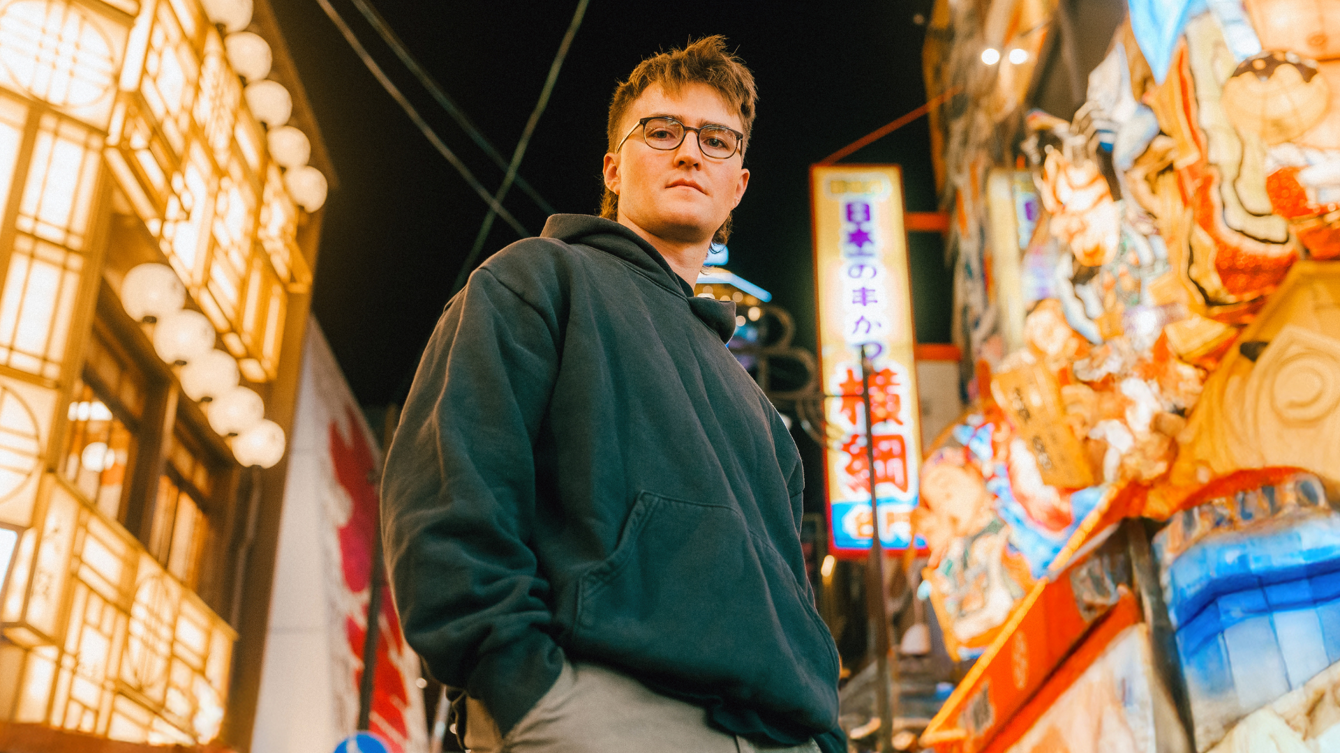 Young man with glasses and a hoodie standing at night in a city area with bright illuminated signs and a colorful festival float or display behind him.