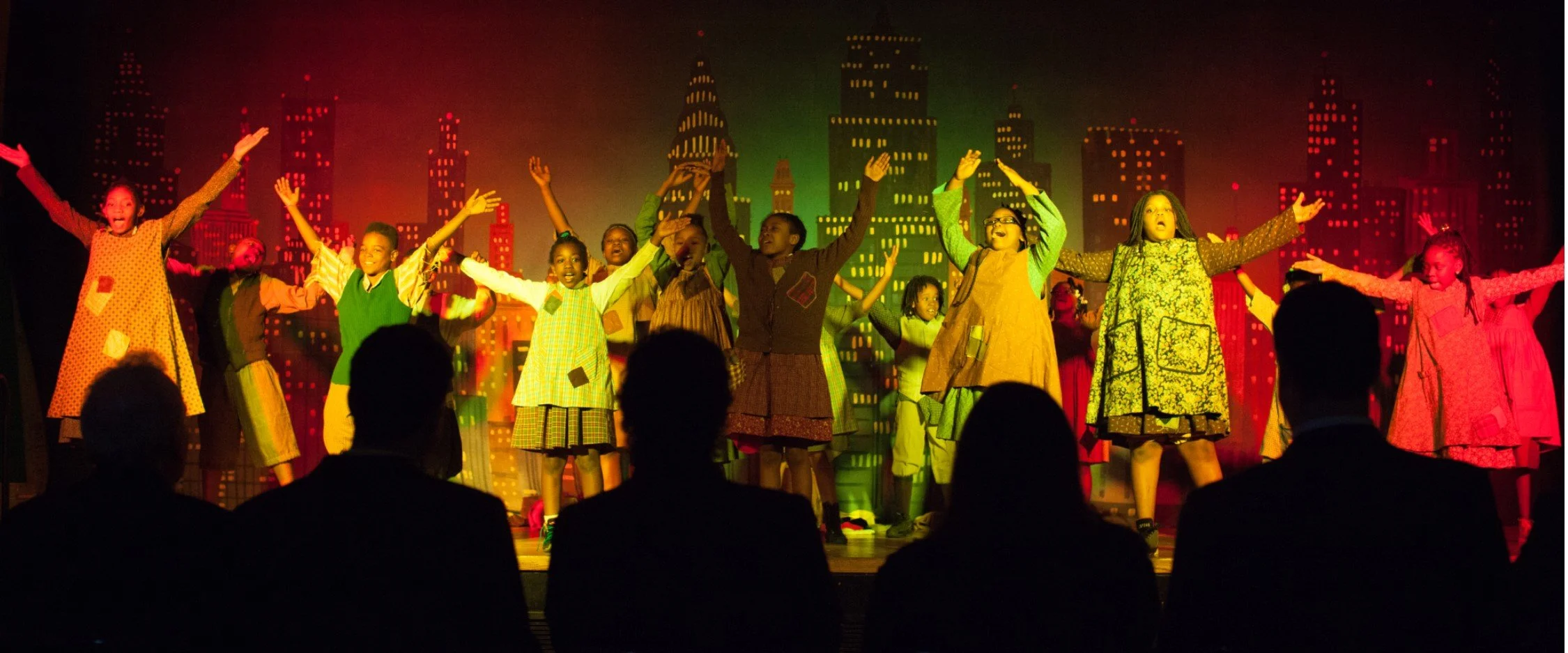 Children performing on stage in a cityscape backdrop with colorful lights, audience watching in silhouette.