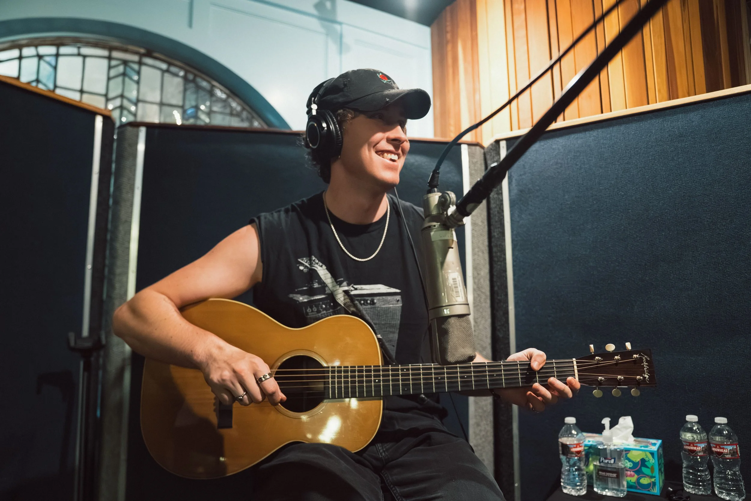 A musician wearing a black cap and sleeveless shirt playing an acoustic guitar in a recording studio, with a microphone in front of them and water bottles on a table nearby.