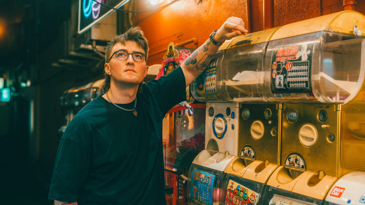 A person with glasses, tattoos, and a chain necklace is reaching into a yellow capsule toy vending machine in a dimly lit arcade or street scene.