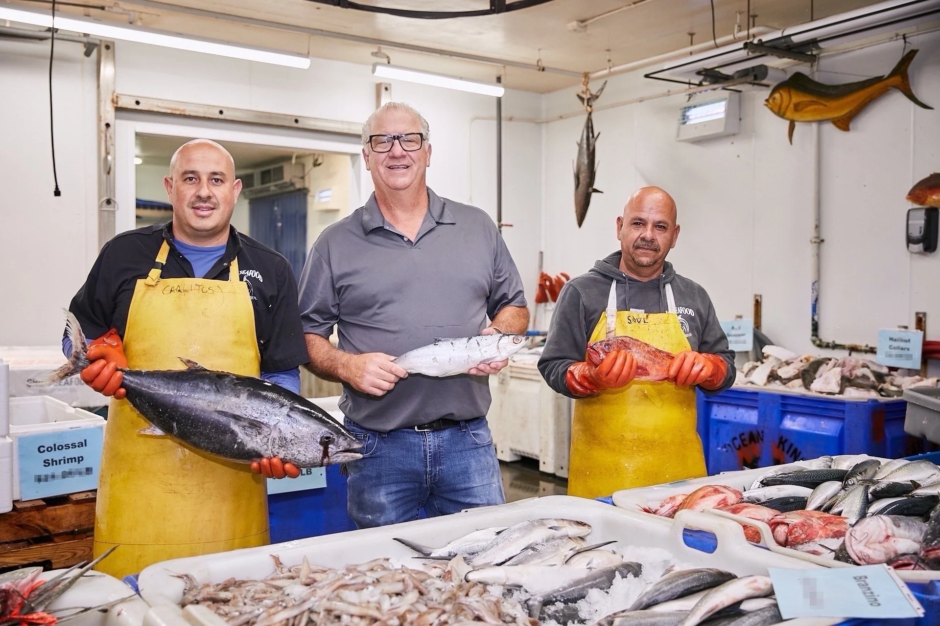 Three men standing behind a seafood display at a fish market, each holding a different type of fish. The man on the left is holding a large fish, the man in the middle is holding a smaller fish, and the man on the right is holding a reddish fish. There are various fish and seafood laid out on the table in front of them, with labels and signs visible in the background.