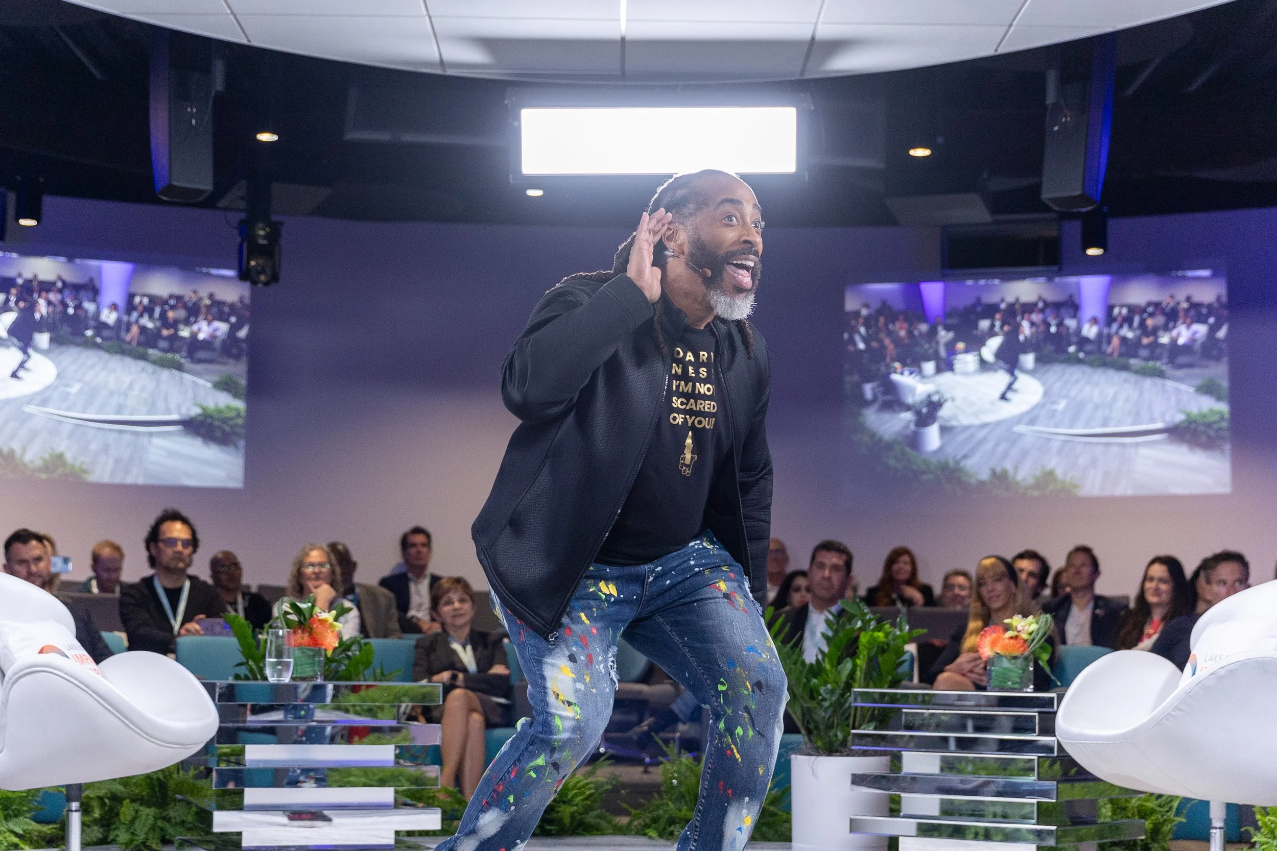 A man with dreadlocks and a beard is speaking or performing in front of an audience, wearing a black jacket, a black T-shirt, and colorful paint-splattered jeans. The audience is seated behind him, and there are two large screens displaying images of a person on a stage. The setting appears to be a conference or event venue with plants and modern chairs.