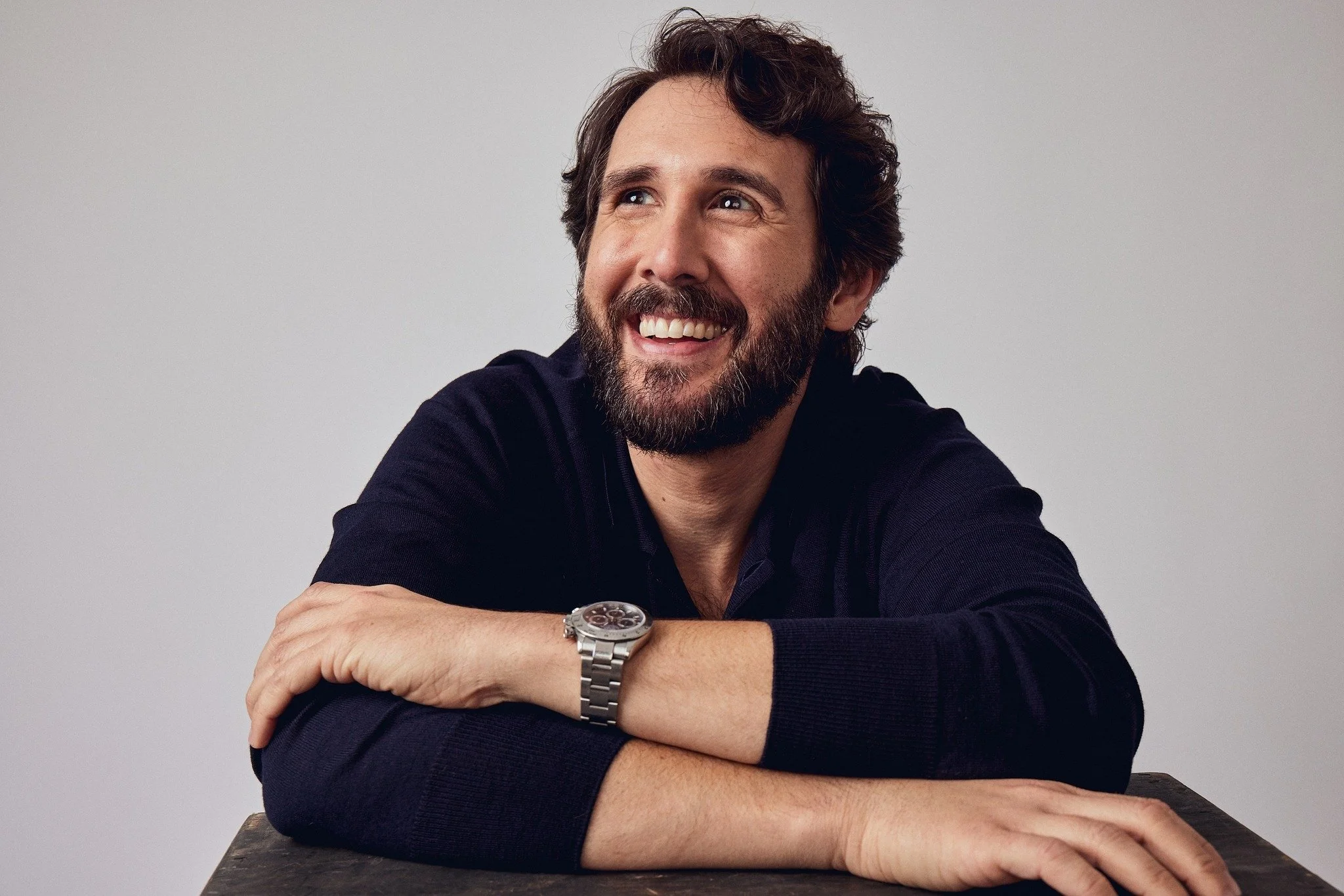 A man with a beard and brown hair smiling and looking away, wearing a black long-sleeve shirt and a silver wristwatch, sitting at a table against a plain white background.