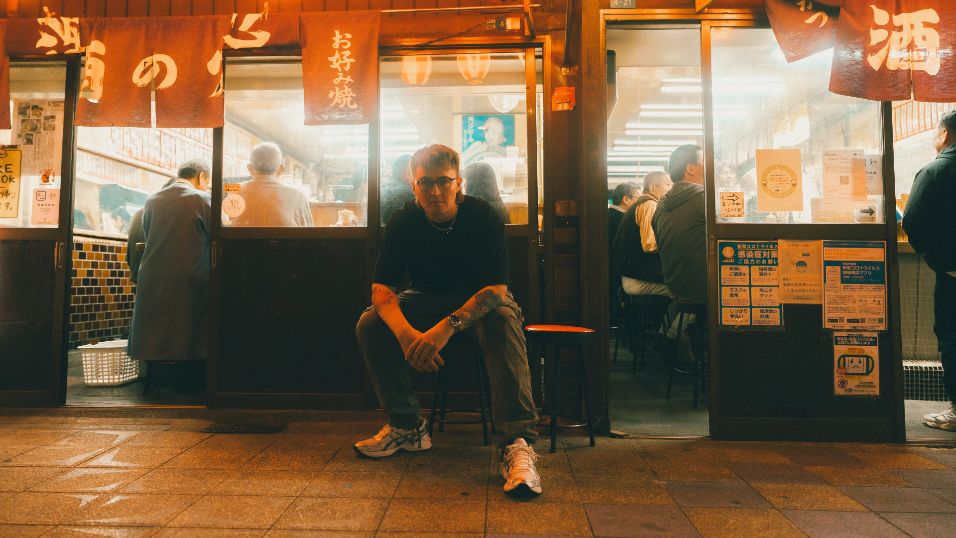 A young man with tattoos, glasses, and wearing casual clothing sits on a stool outside a Japanese restaurant. Inside, people are dining, with red noren curtains hanging above the entrance and warm lighting.