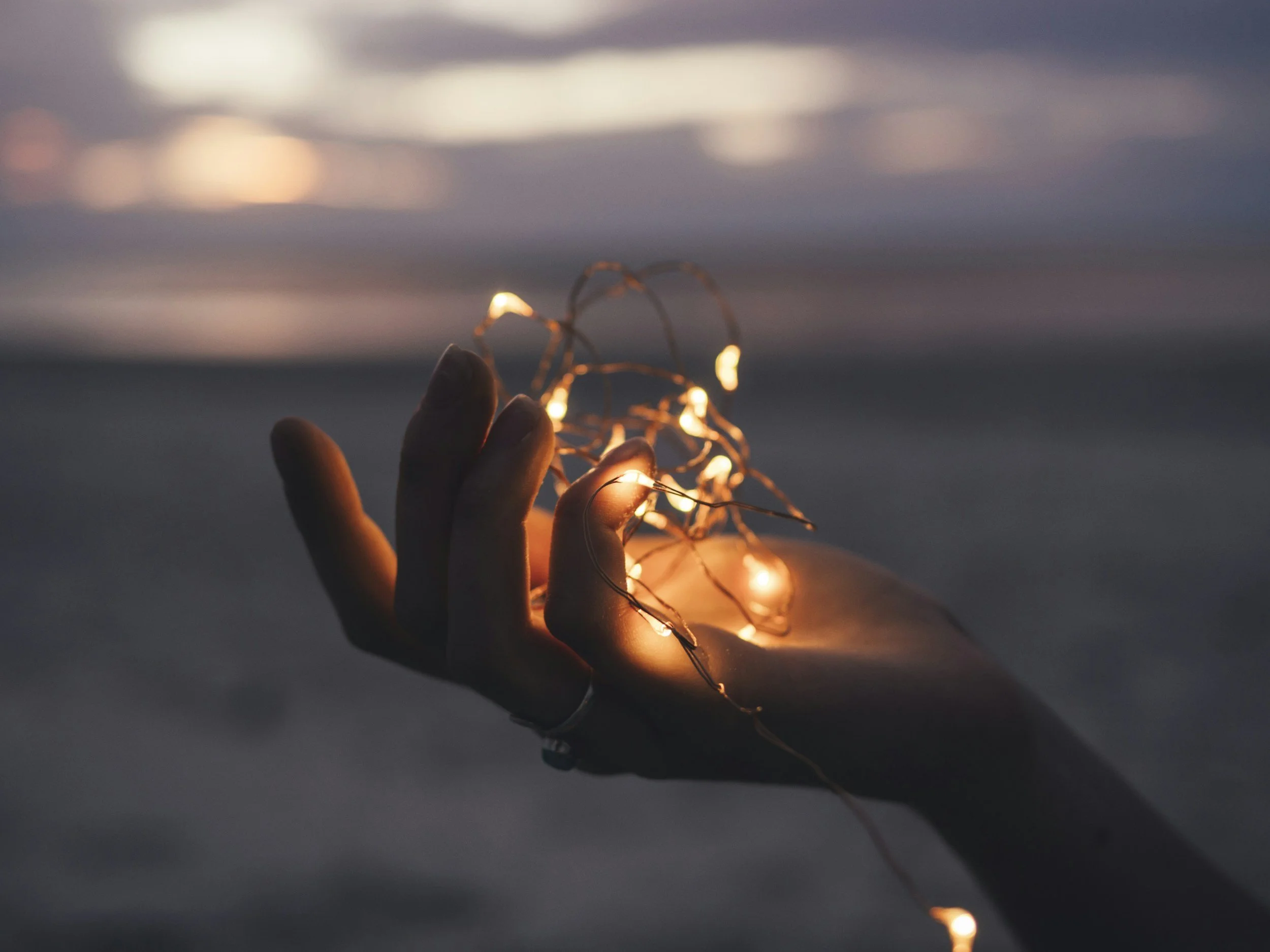 A person's hand holds a string of small glowing fairy lights against a blurred, dusky sky background.