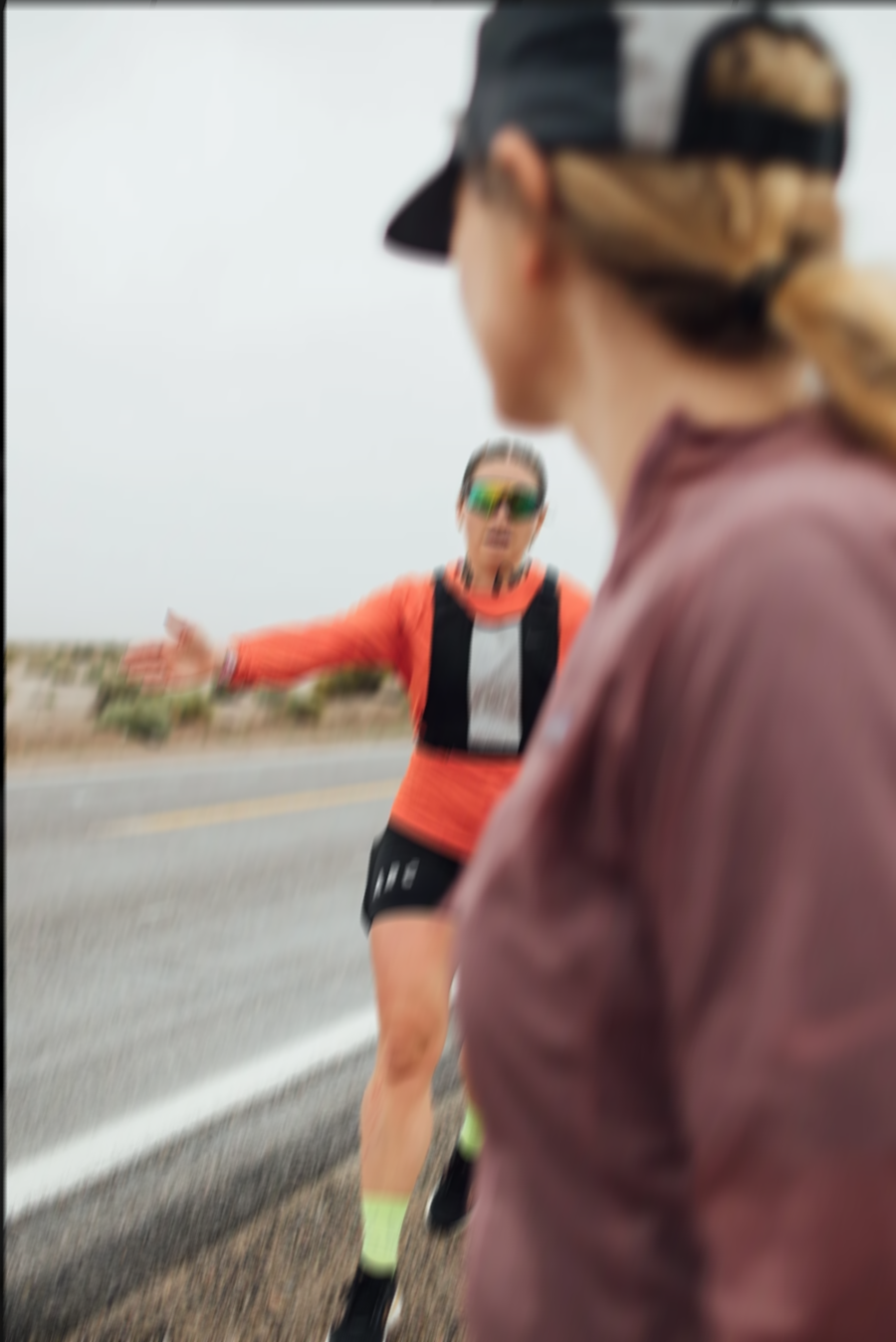 A person in running gear being approached or stopped by a woman in a cap on a road with an overcast sky.