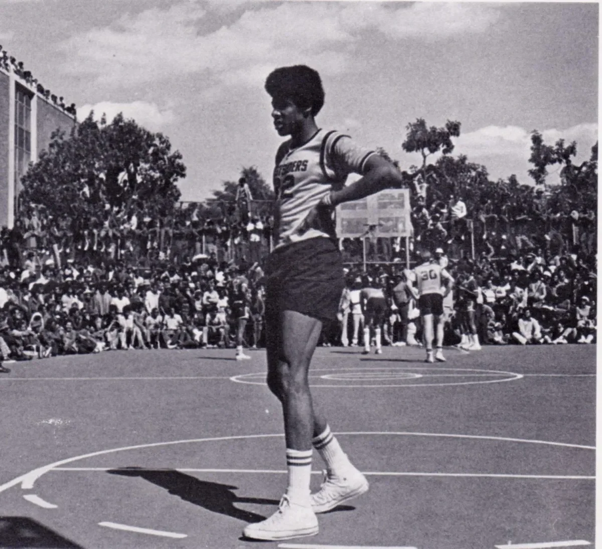 Black and white photo of a basketball player standing on the court during a game with a crowd of spectators seated on bleachers and trees in the background.