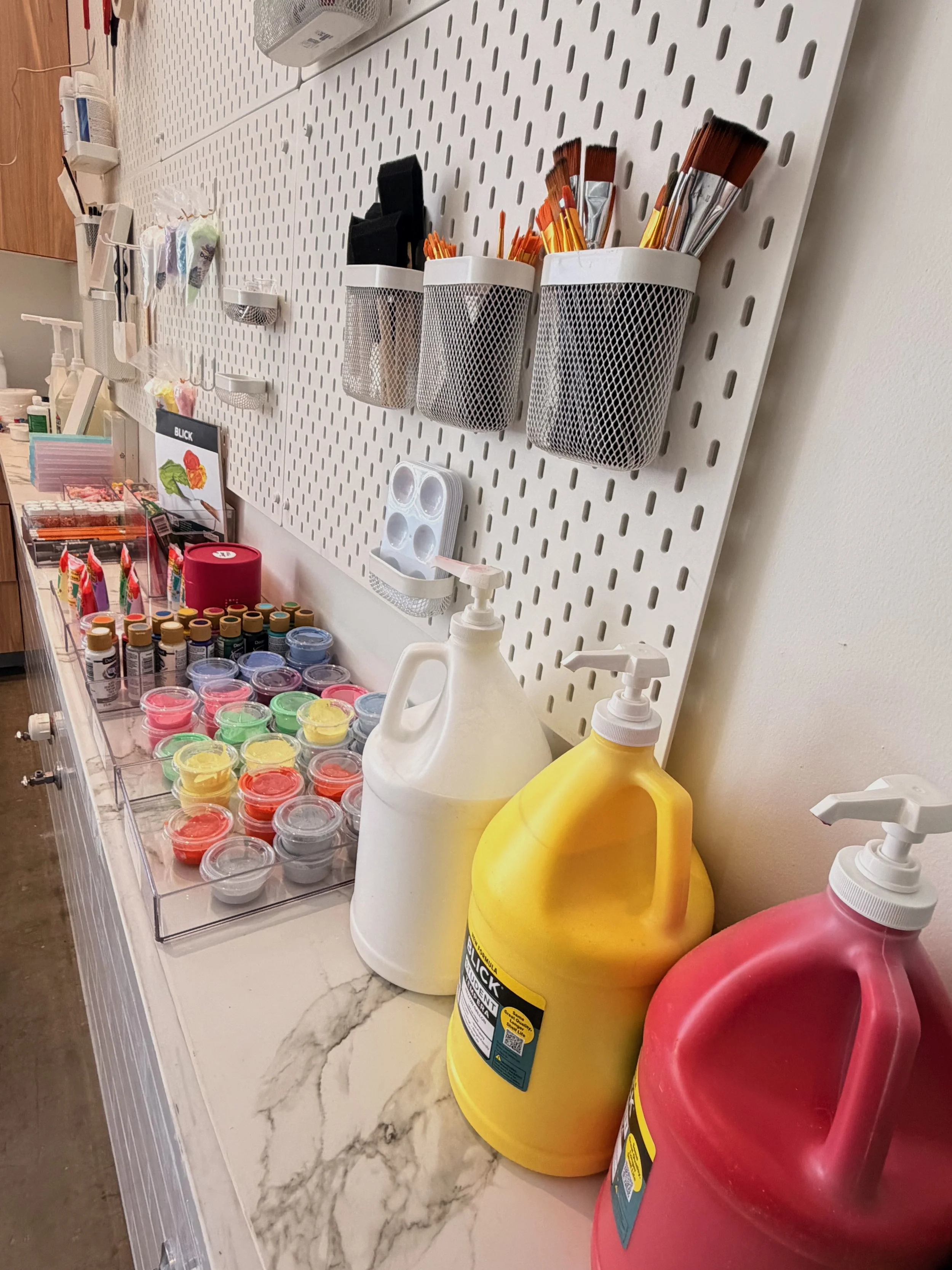 Counter with various art supplies including paints, brushes, and containers, with cleaning supplies in the foreground and a pegboard with small storage cups behind.