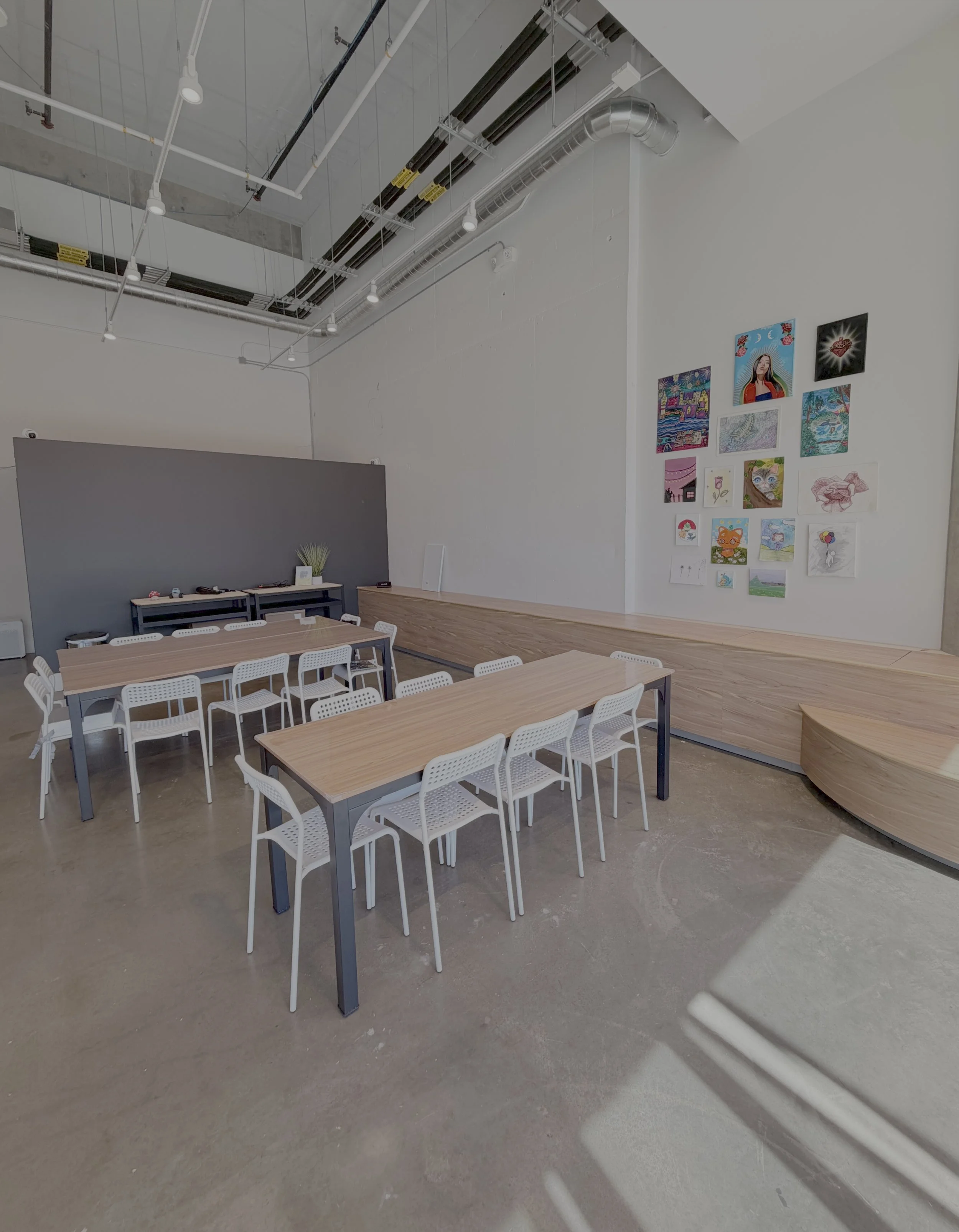 Modern indoor space with wooden tables and white chairs, decorative artwork on the wall, and industrial ceiling with exposed pipes.