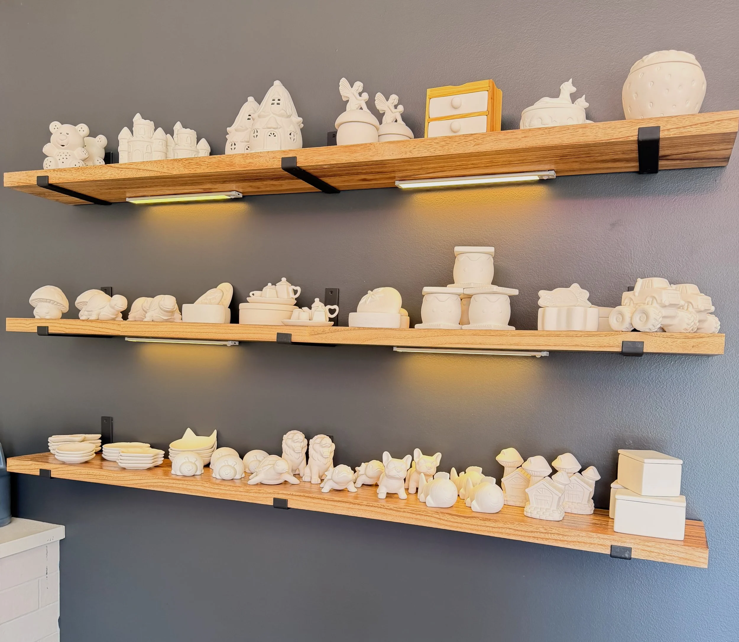 Three wooden shelves mounted on a gray wall, displaying various white ceramic figurines and decorative objects, including animals, buildings, and household items, with under-shelf lighting.