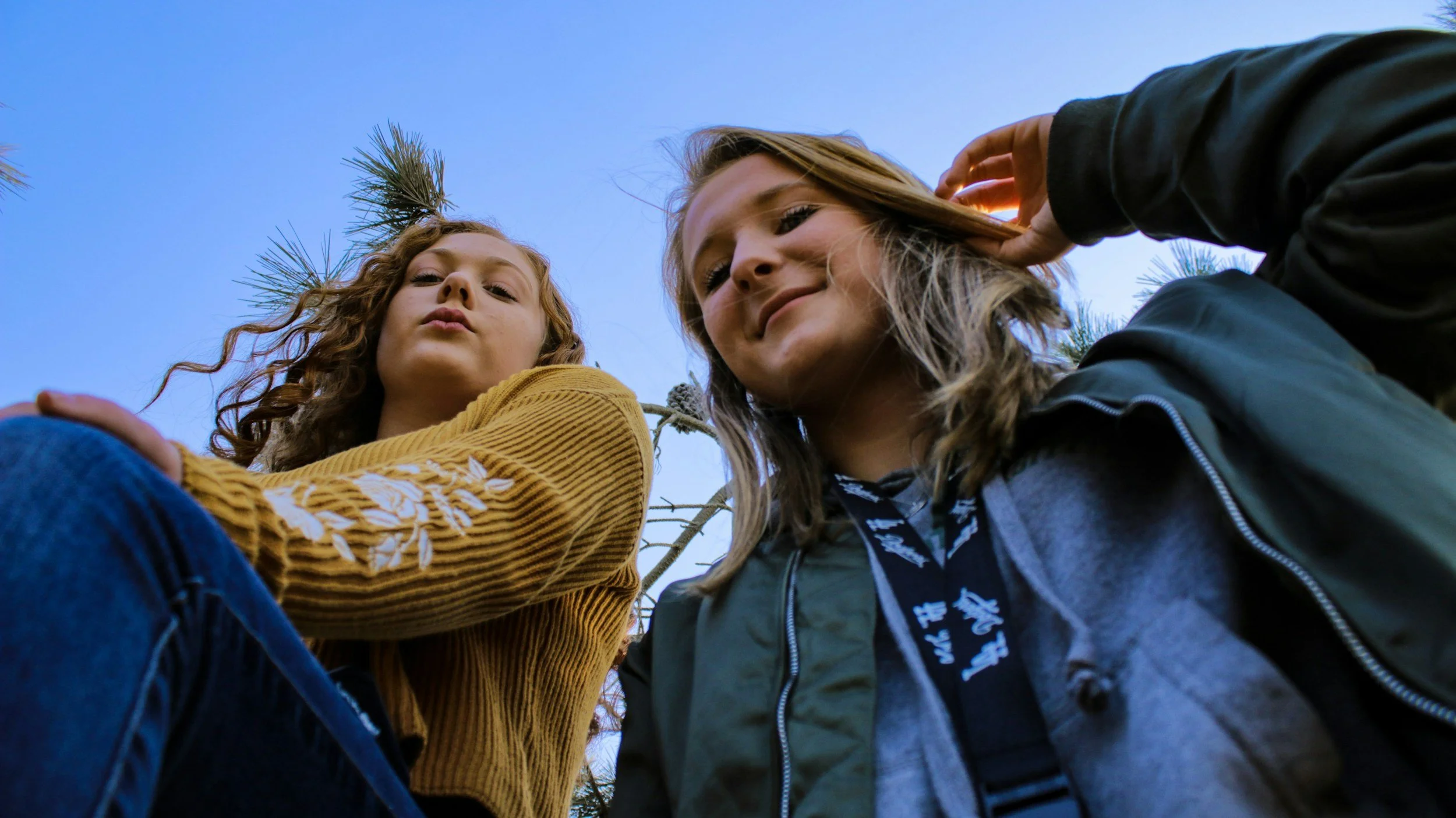 Two young women looking down at the camera, standing outdoors with pine trees and a blue sky in the background.