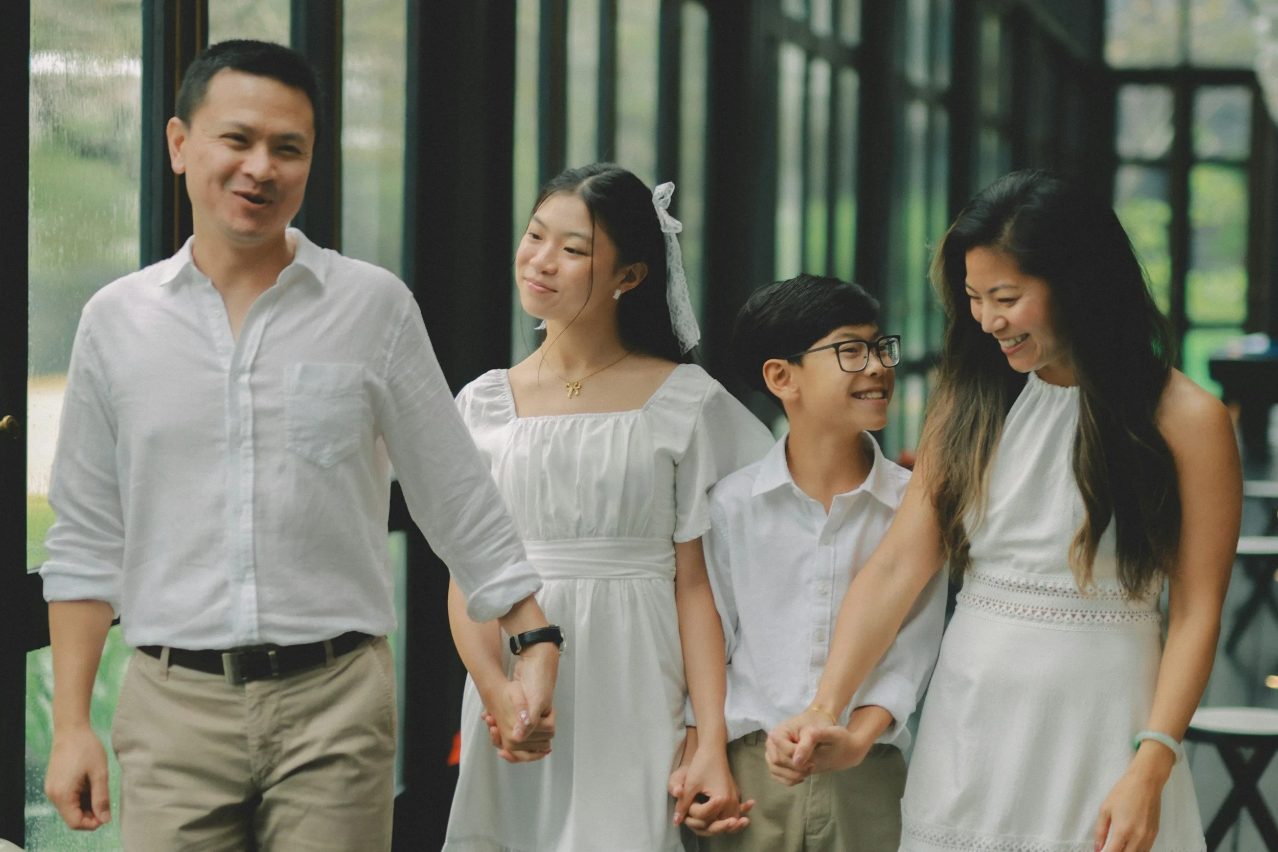 A family of five holding hands, smiling, walking indoors near large windows with greenery outside.