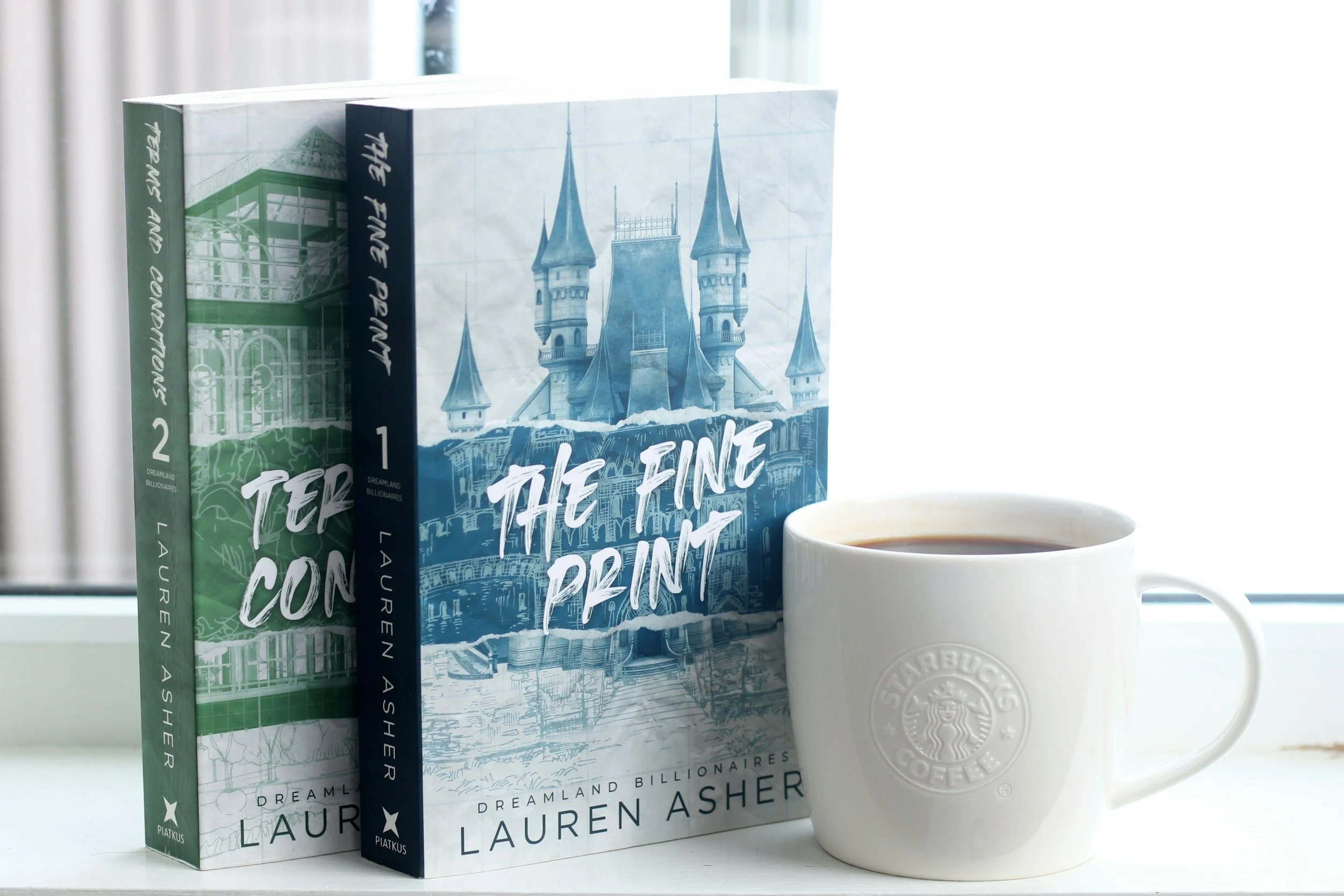 Two copies of the book "The Fine Print" by Lauren Asher and a white Starbucks coffee mug filled with coffee placed on a white surface in front of a window.