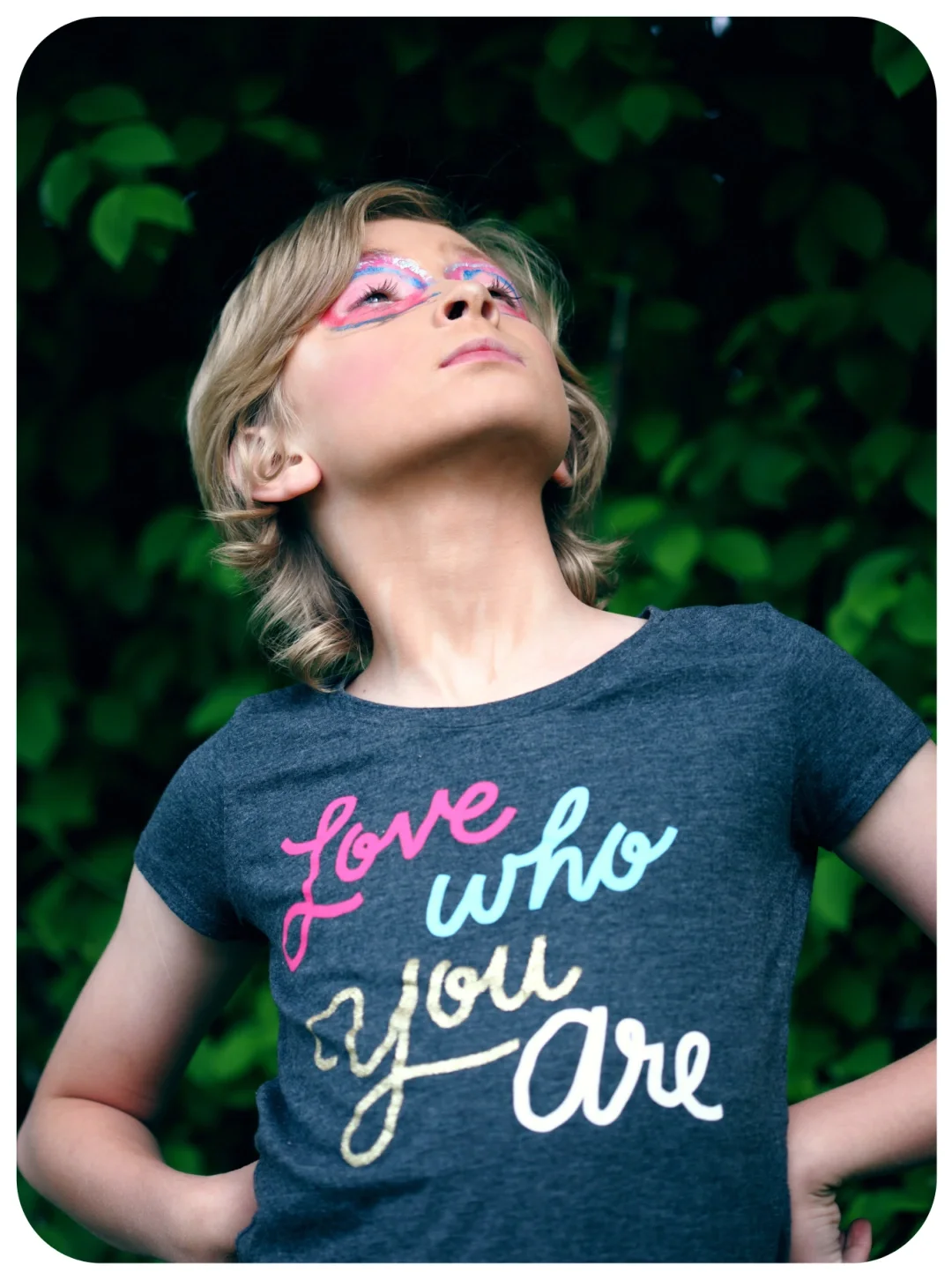 Person with colorful face paint looking upward outdoors, wearing a dark gray t-shirt with the words 'love who you are' in colorful cursive.