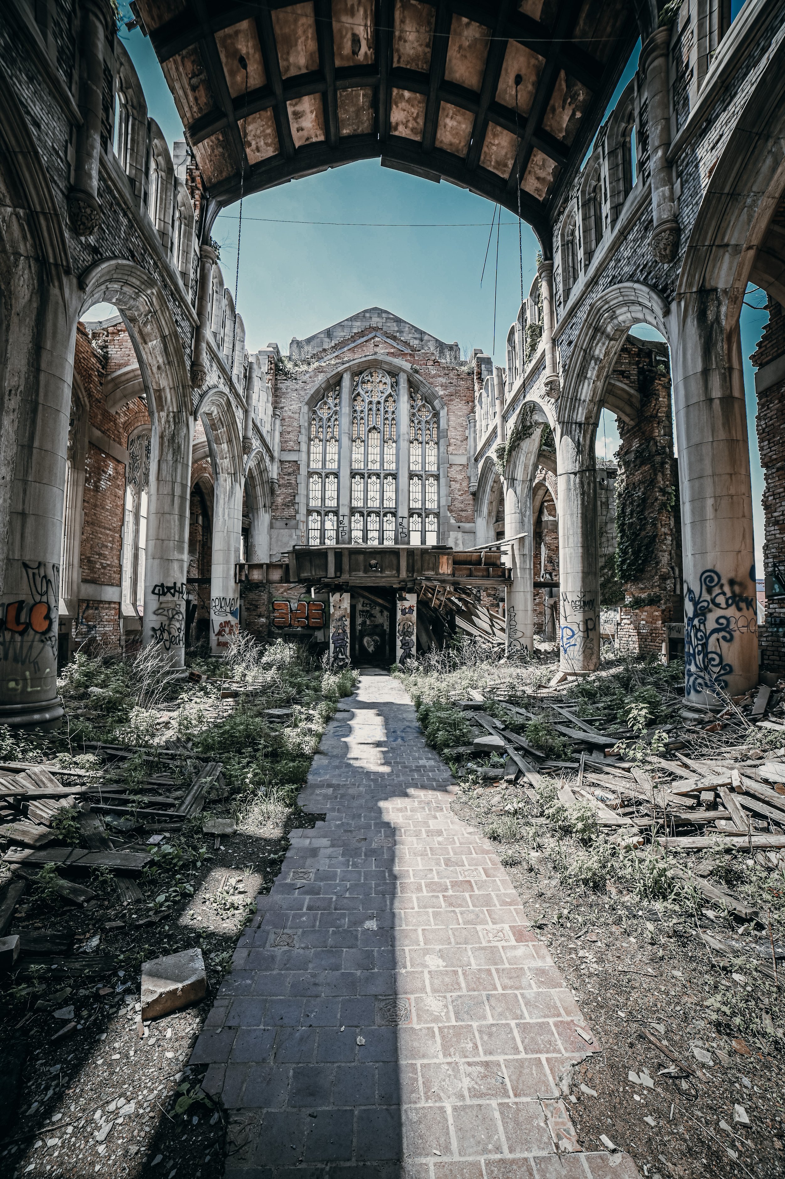 An abandoned, partially ruined church with broken windows and graffiti on the columns. Overgrown plants and debris line the brick pathway leading to the altar area.