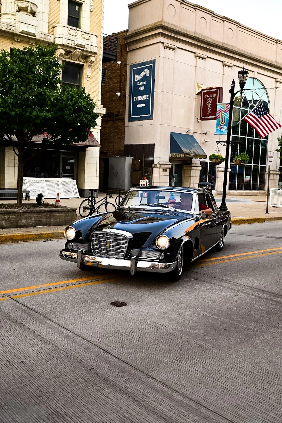 A vintage black car driving down a city street with American flags hanging from a lamppost. Buildings and a sign for a dance hall are visible in the background.