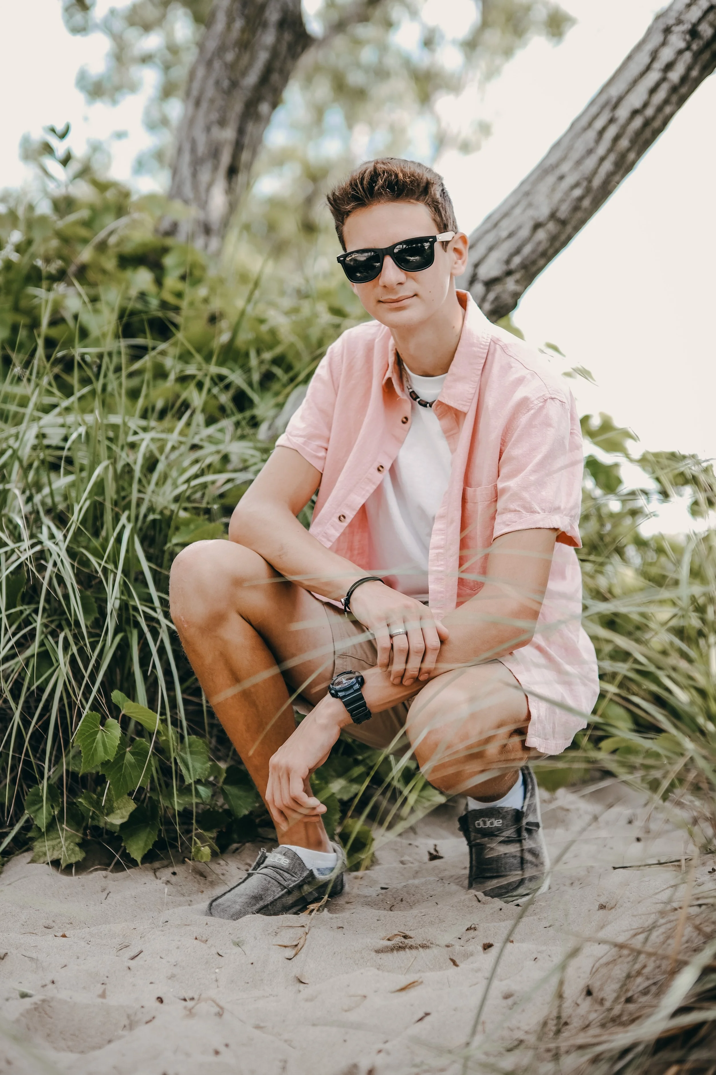 A young man with short dark hair, wearing sunglasses, a light pink short-sleeve shirt over a white T-shirt, beige shorts, and sneakers, crouching on sandy beach surrounded by green foliage and trees.