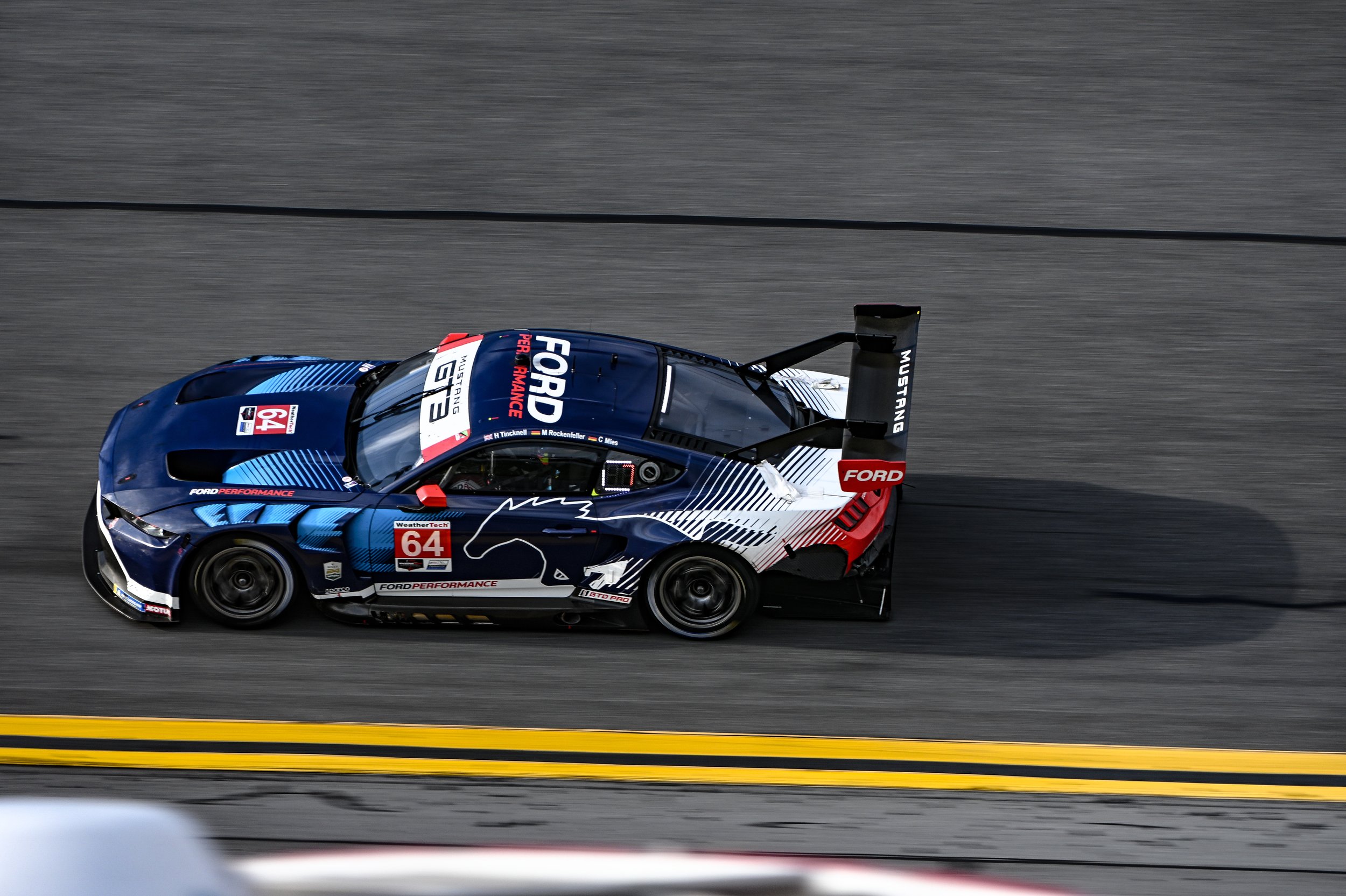 A blue, white, and red Ford race car with the number 64 on the side, driving on a race track with yellow lines, viewed from above.