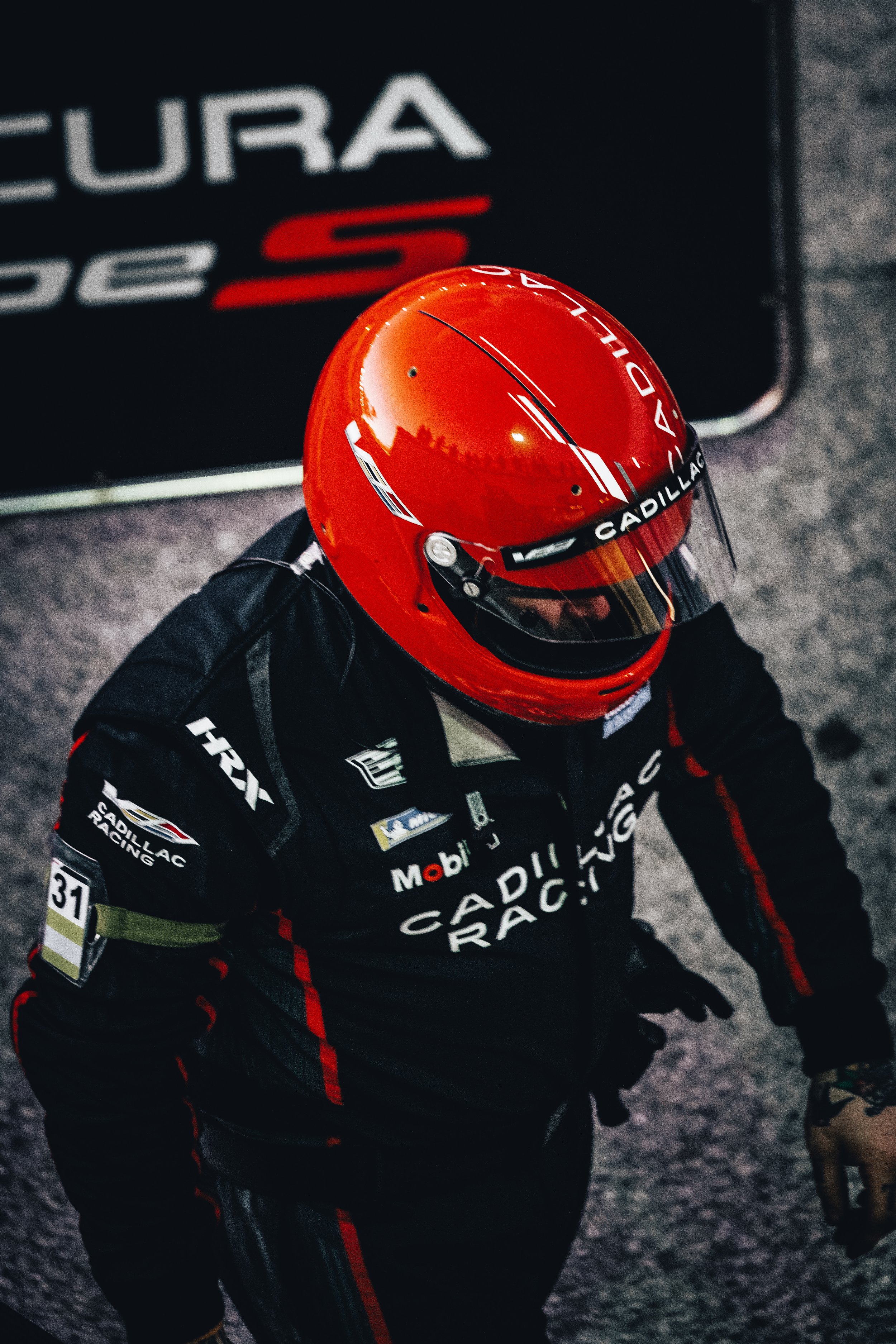 Race car driver in black racing suit with Cadillac Racing and Mobil logos, wearing a red helmet with a tinted visor, standing on a gray race track surface.