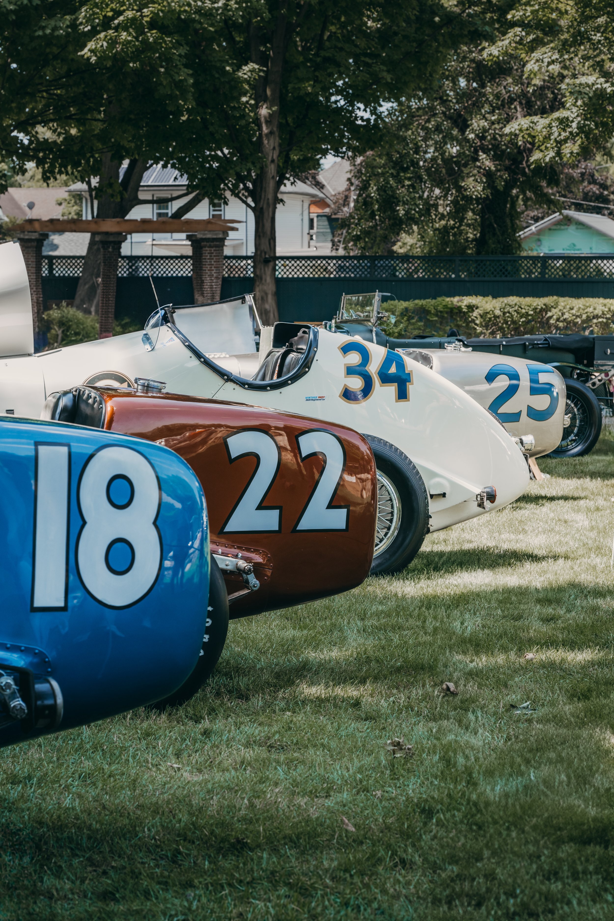 Multiple vintage race cars with racing numbers parked on a grassy area under trees in a backyard setting.