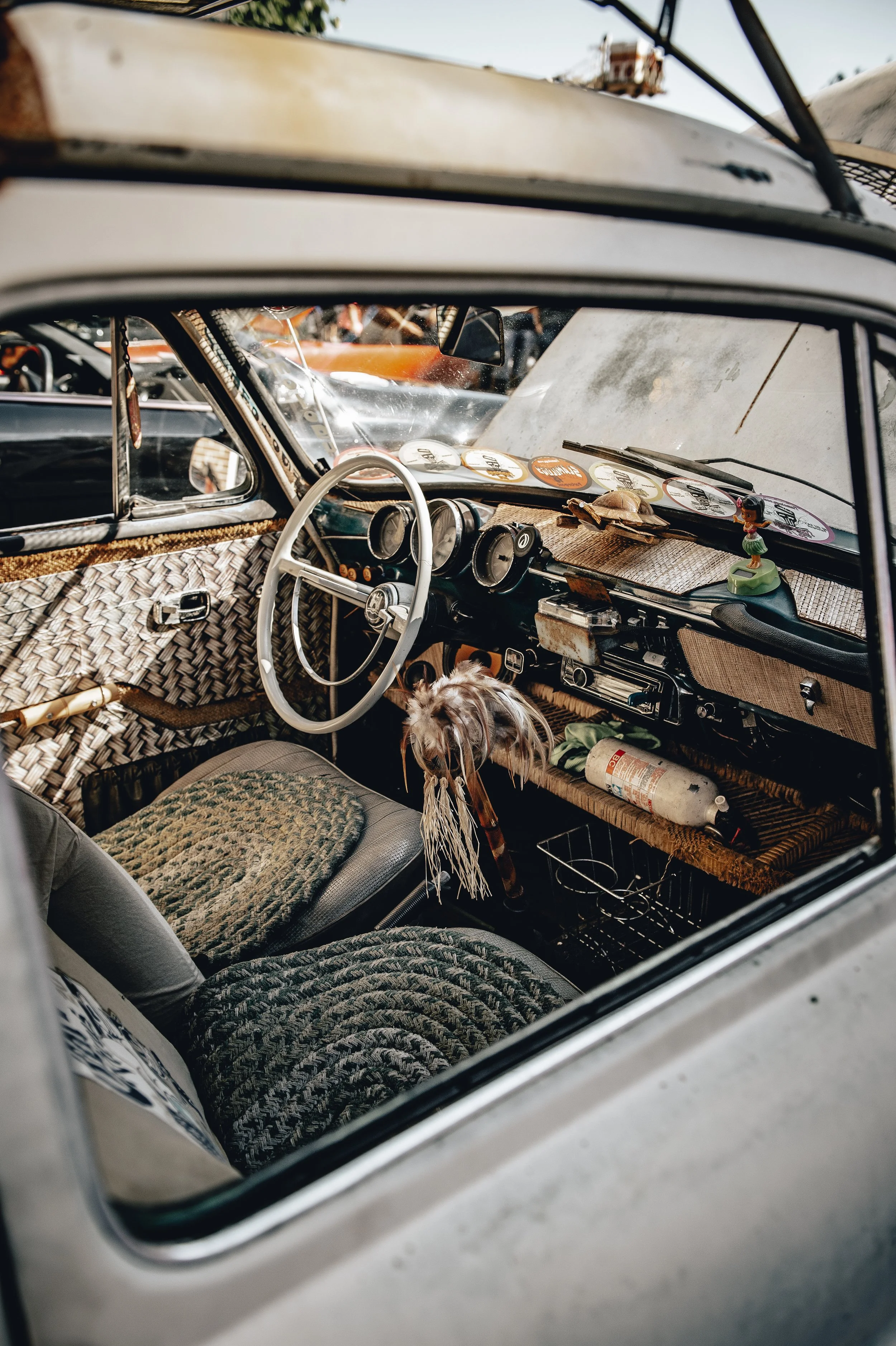 Interior of an old, vintage car with woven fabric dashboard and seat covers, featuring a feathered dreamcatcher hanging from the steering wheel, and a fire extinguisher on the passenger side.