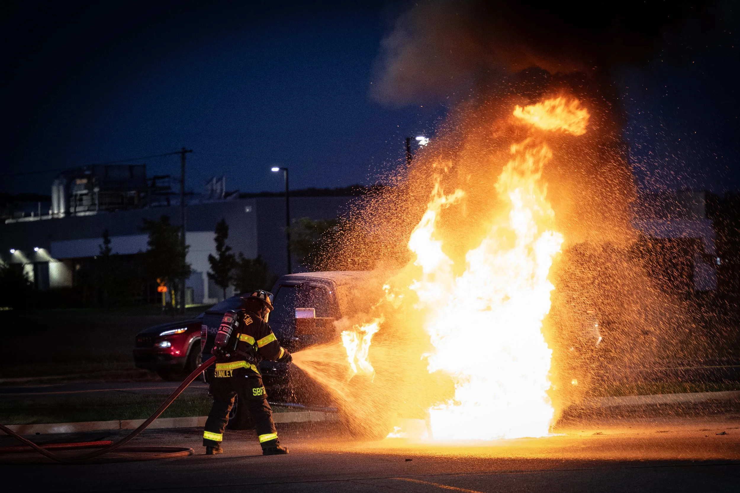 Firefighter fighting a car fire at night with a hose in an urban area.