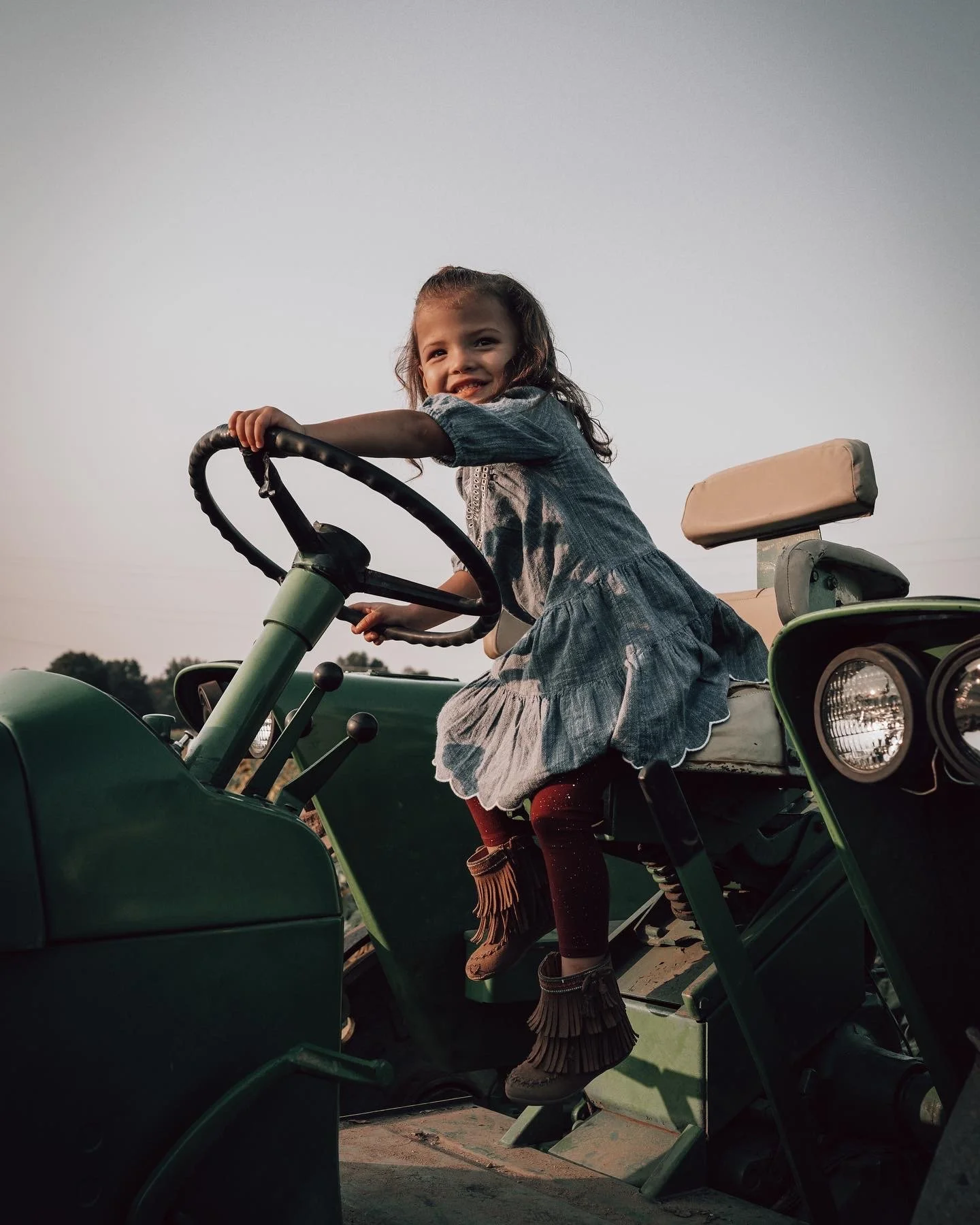 A young girl dressed in a denim dress and fringed boots sitting on a green tractor, smiling at the camera during sunset.