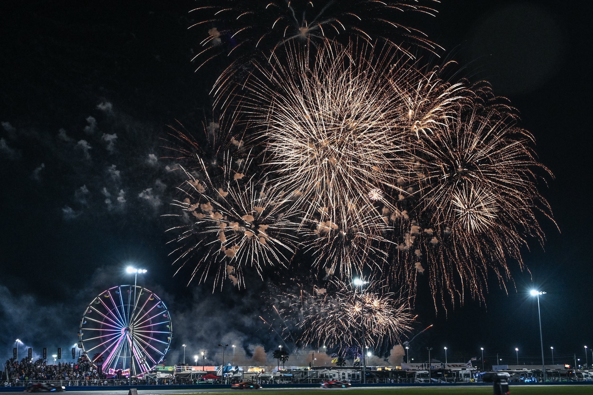Night sky lit up with fireworks at a nighttime fair or carnival, with a ferris wheel illuminated with colorful lights and a crowd of people gathered below.