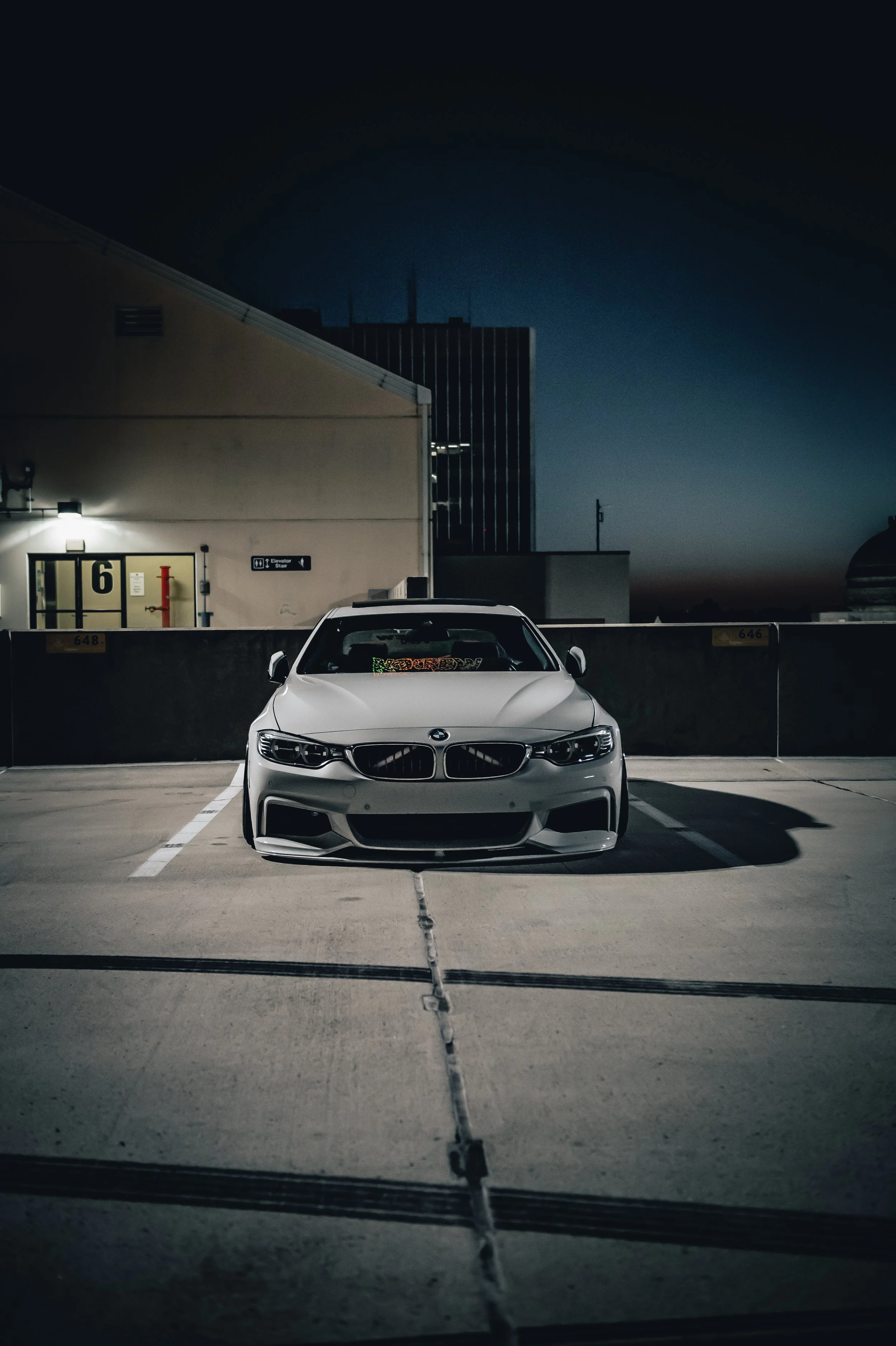 A white BMW parked alone in an empty rooftop parking lot at night, with a dark cityscape and building in the background.