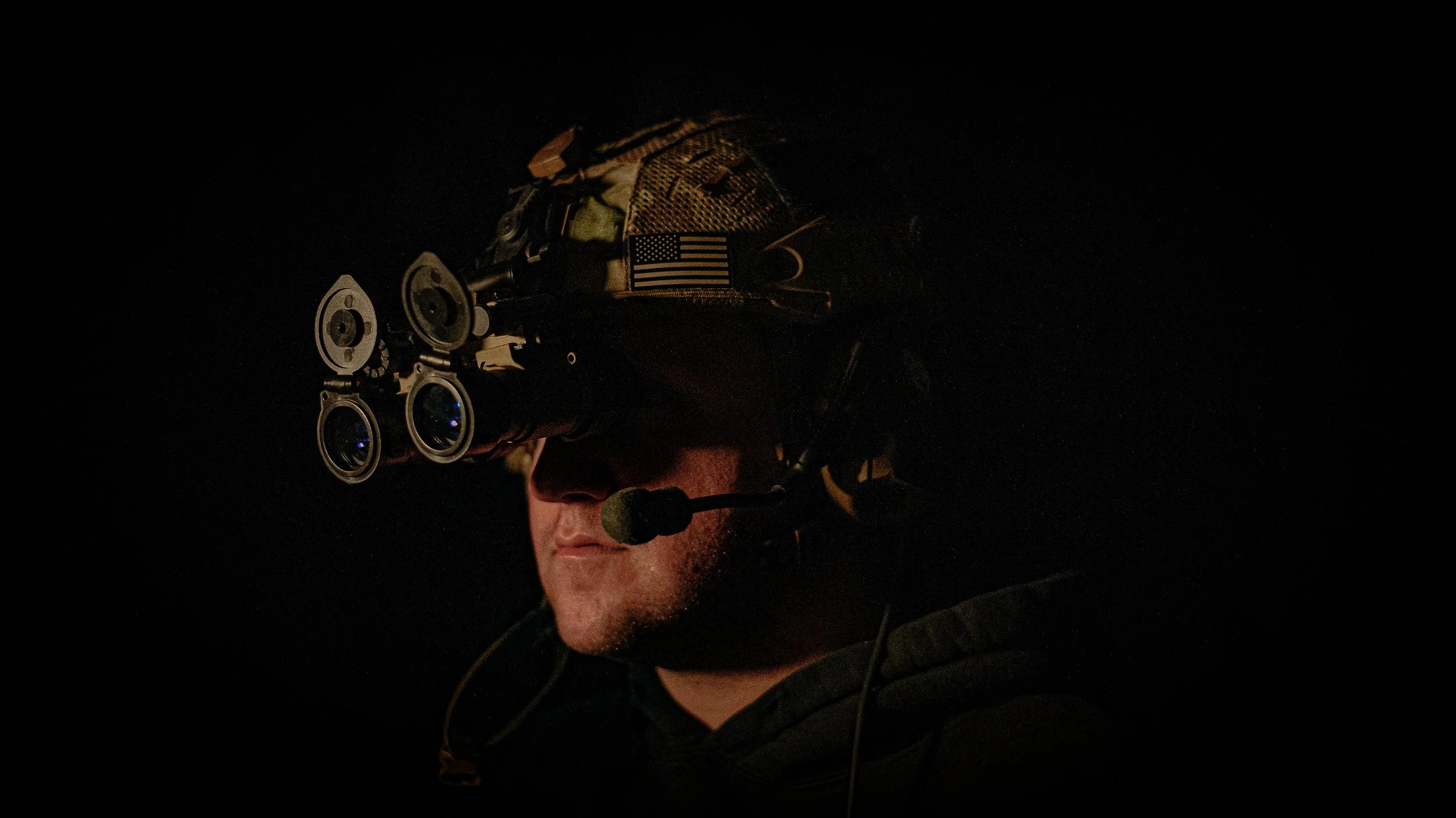 A soldier wearing night vision goggles and a camouflage helmet with an American flag patch, speaking into a microphone in a dark setting.