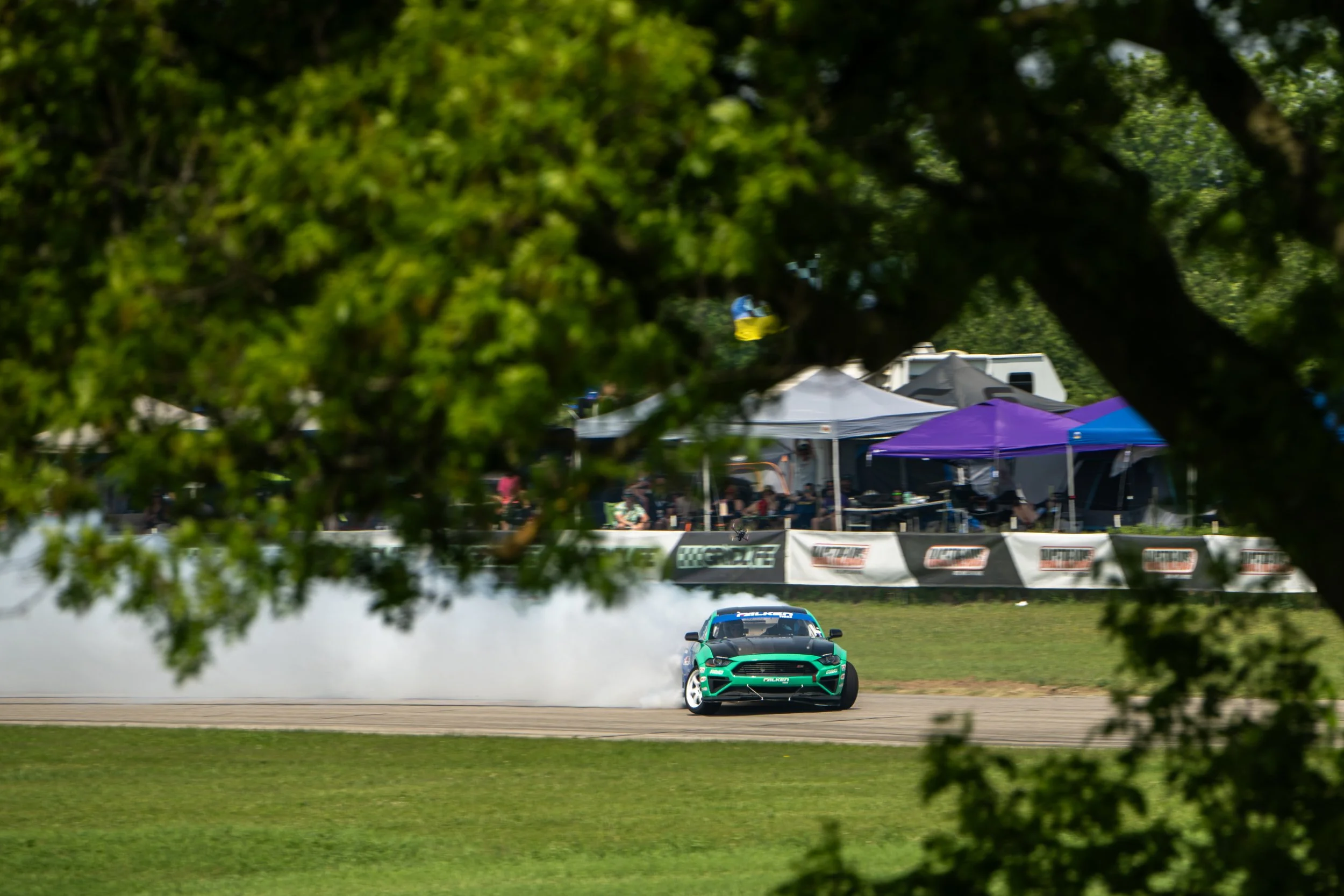 A green and black race car drifting on a race track, producing smoke, with tents and spectators in the background, partially framed by tree branches.
