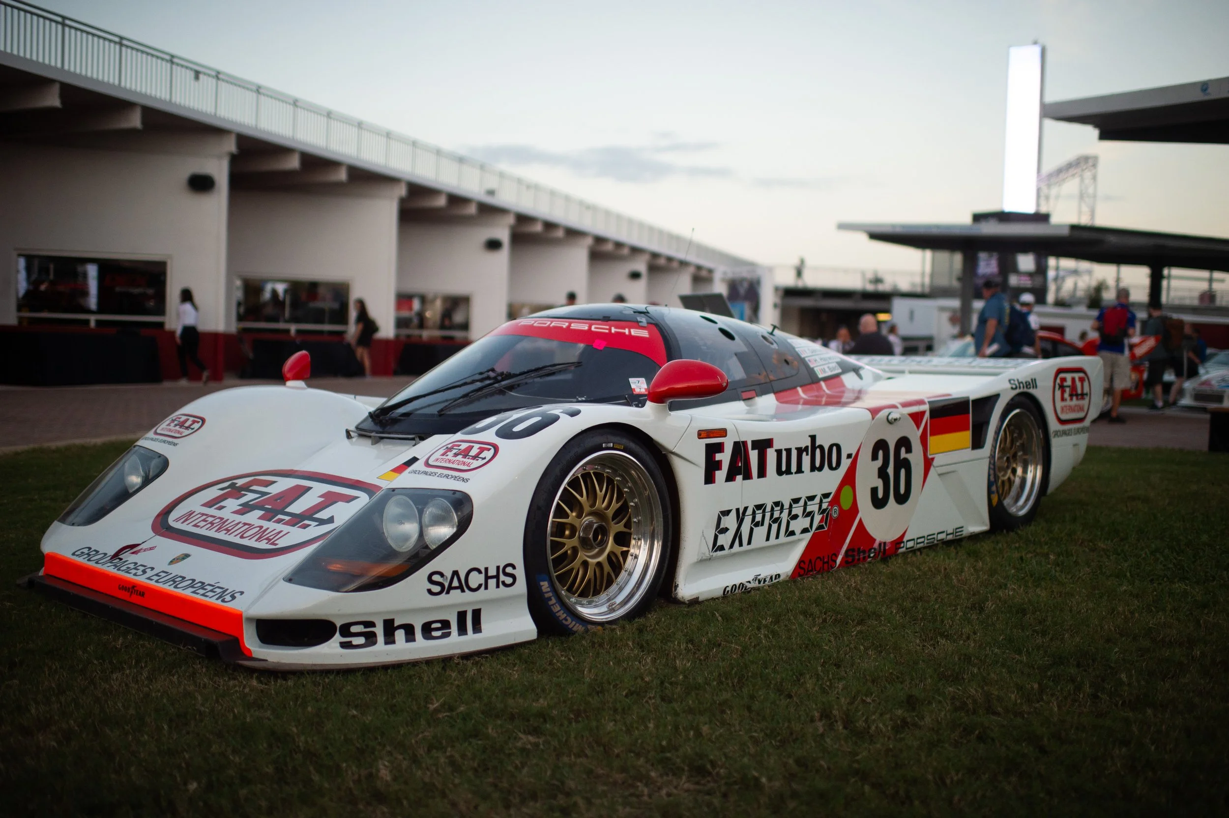 A Porsche race car with the number 36, featuring various sponsor logos including FAT, Shell, Sachs, and EAT International, parked on grass at an outdoor event during dusk.