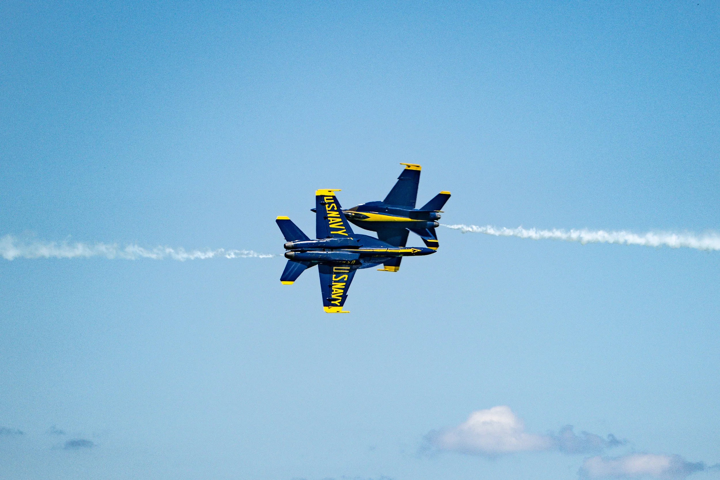 Two U.S. Navy Blue Angels fighter jets flying upside down in formation with white smoke trails against a blue sky.
