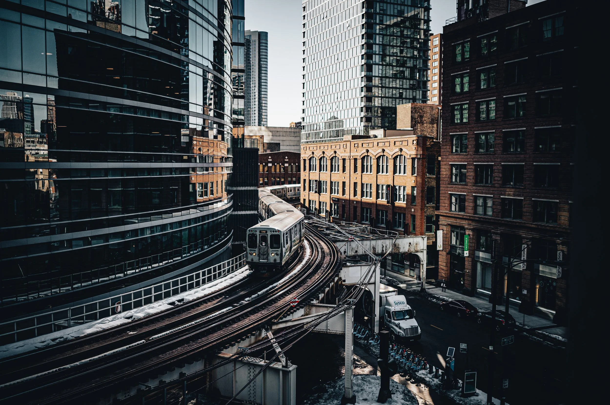 Cityscape with modern glass buildings, a train on an elevated track, and snow on the ground and tracks.